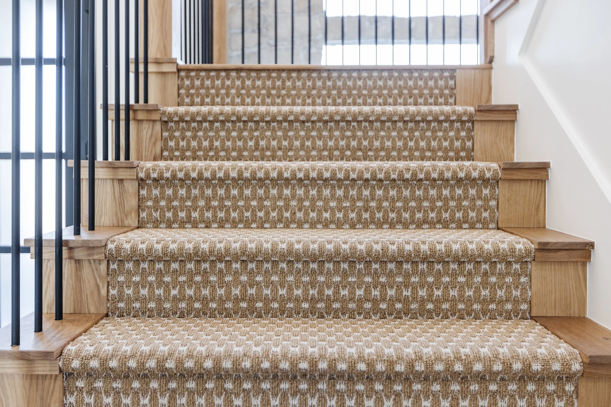 Wooden staircase with patterned carpeted steps leading upward, flanked by black metal railing on the left and a white wall on the right, illuminated by natural light from a window at the top.