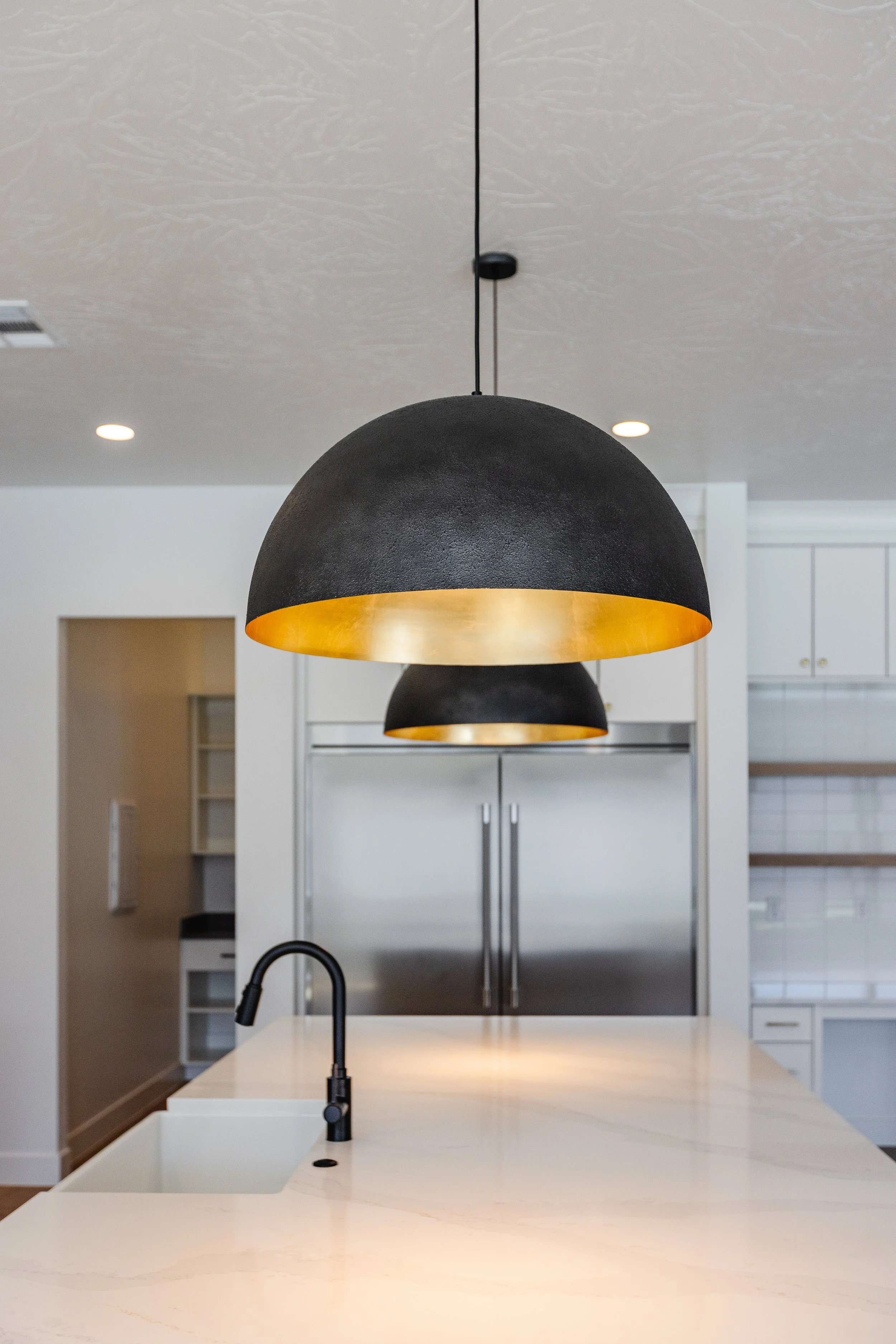 Pendant light fixture with a black exterior and gold interior hangs over a kitchen island with a black faucet, in a modern kitchen with white cabinetry and tiled backsplash.