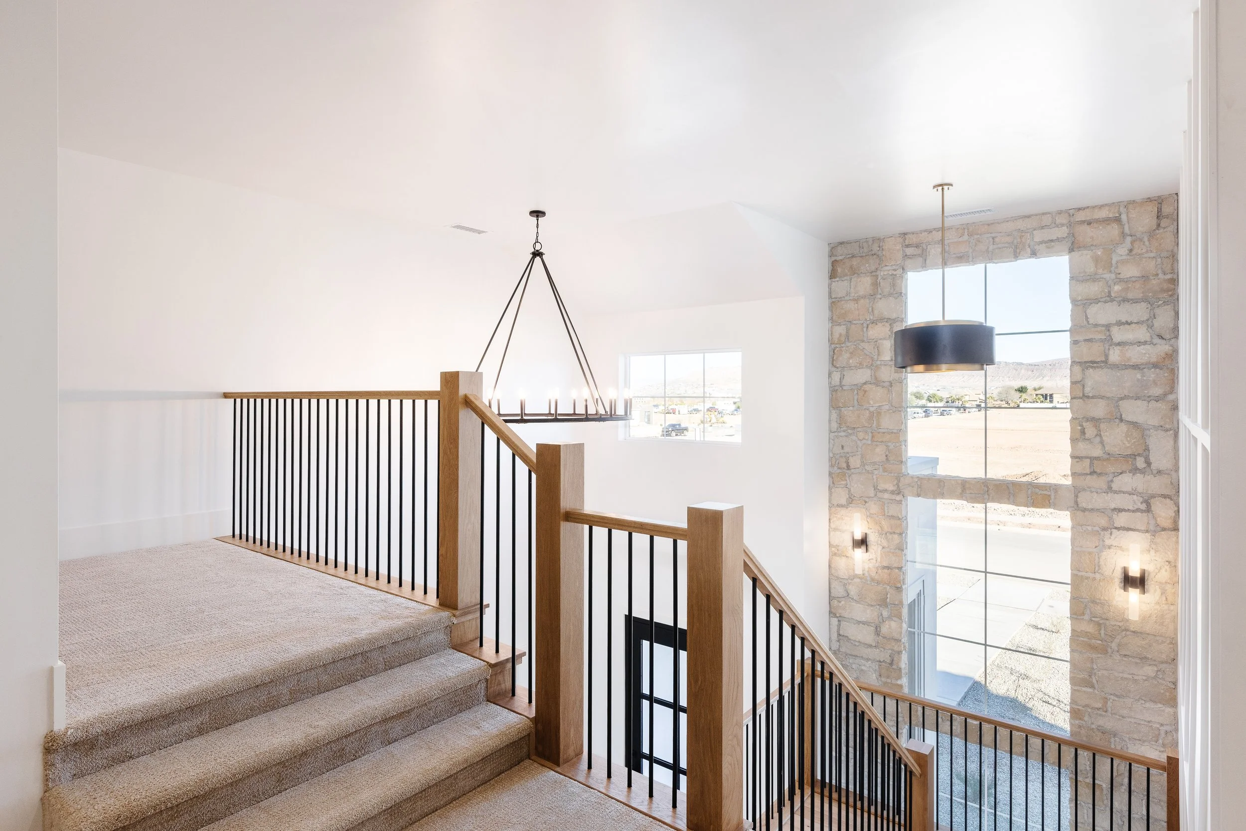 Interior view of a staircase with beige carpet, black metal railing with wooden handrail, a large window, stone accent wall, and hanging light fixtures.