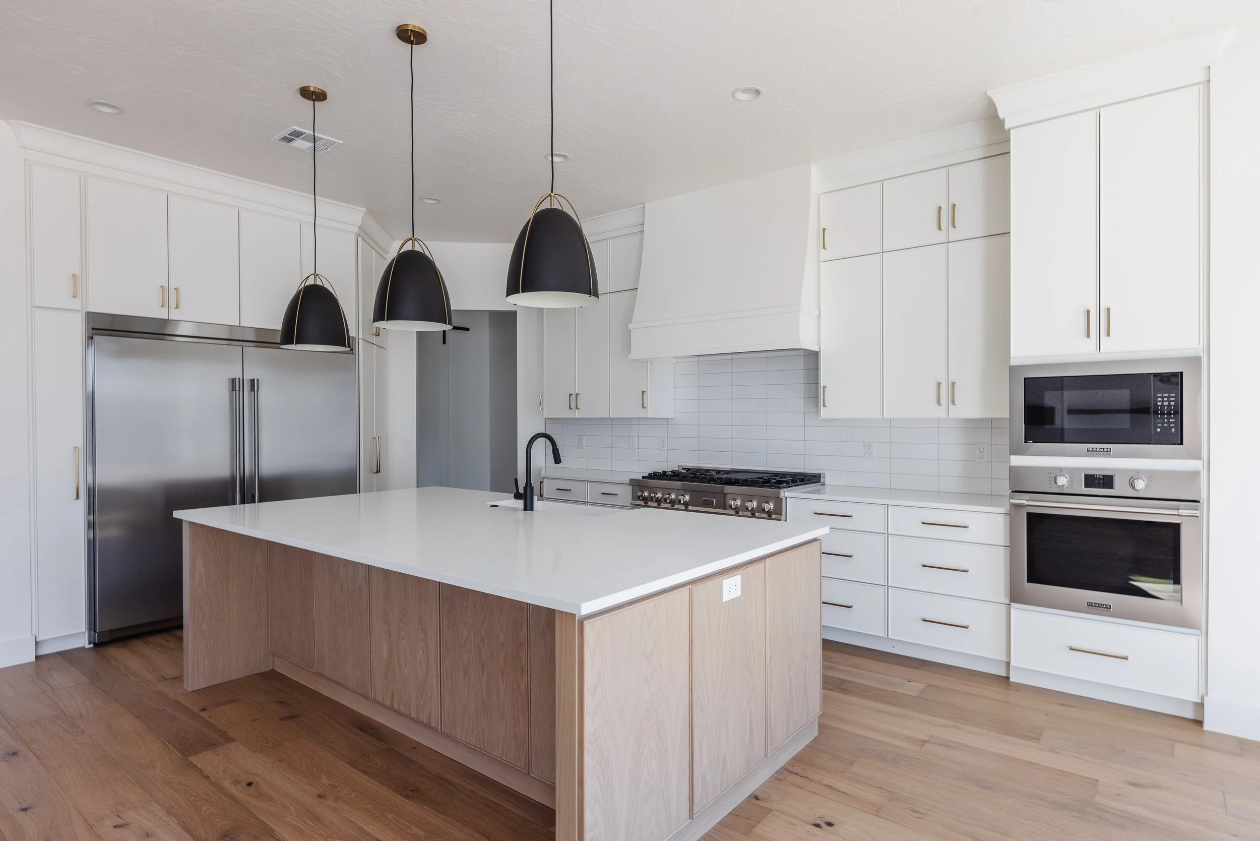 Modern kitchen with white cabinets, a large island with a light wood base and white countertop, black pendant lights, stainless steel refrigerator, built-in oven and microwave, and a gas stove with white backsplash.