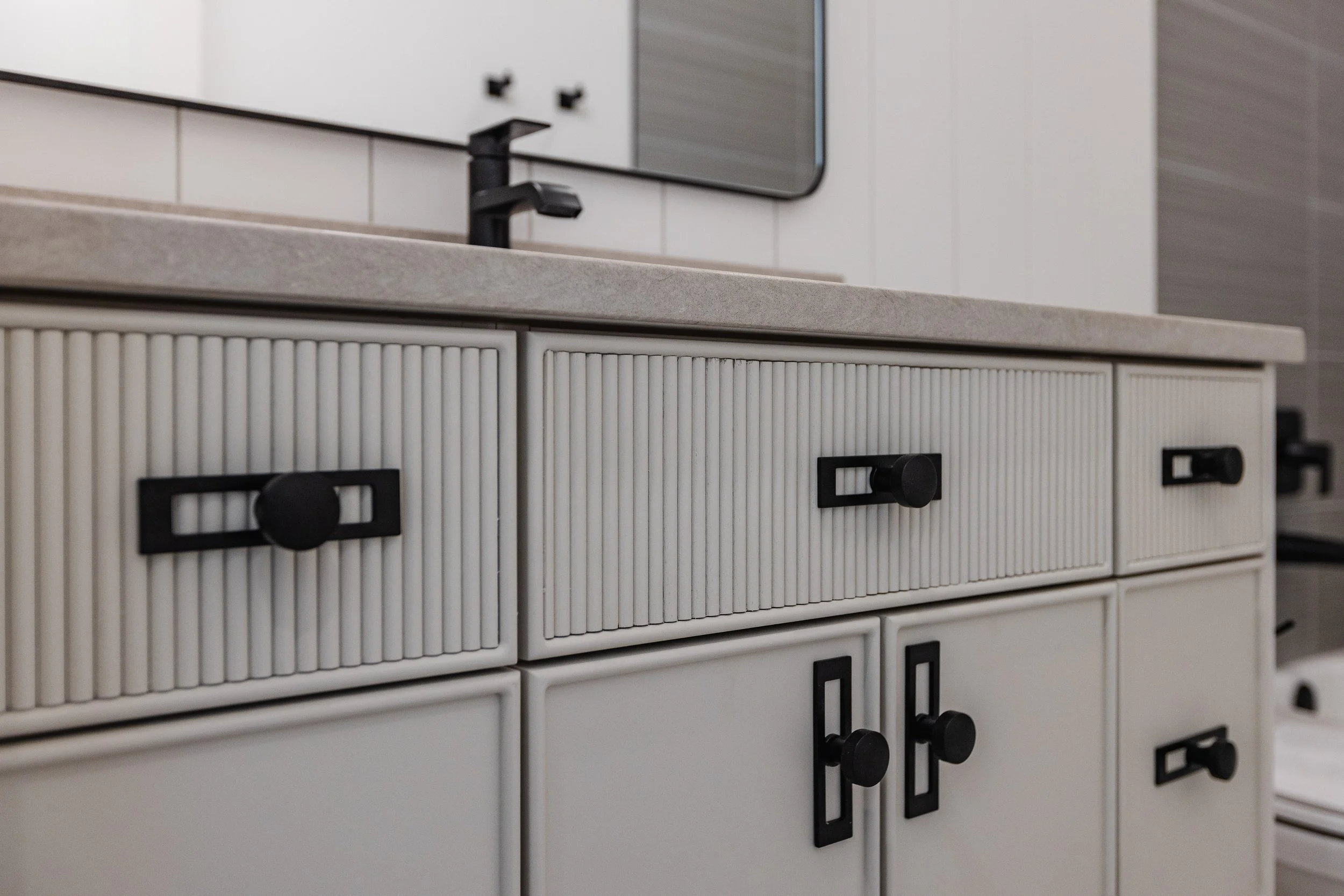 Close-up of a white kitchen cabinet with black knobs, a beige countertop, and a black faucet, with a mirror and tiled wall in the background.