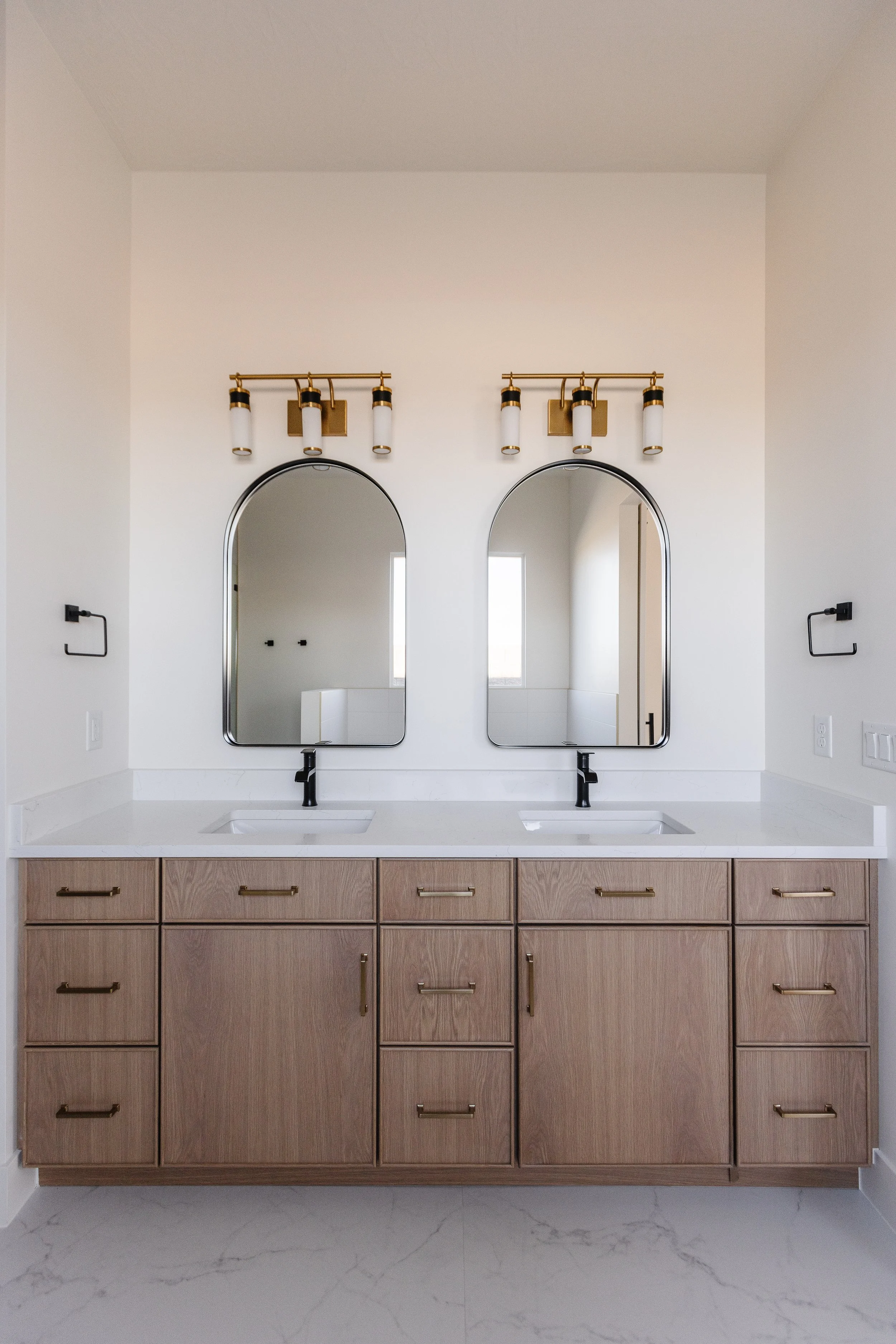 Bathroom vanity with two arched mirrors, black faucets, and a wooden cabinet with multiple drawers, bathroom lighting fixtures above mirrors, white walls, and marble floors.
