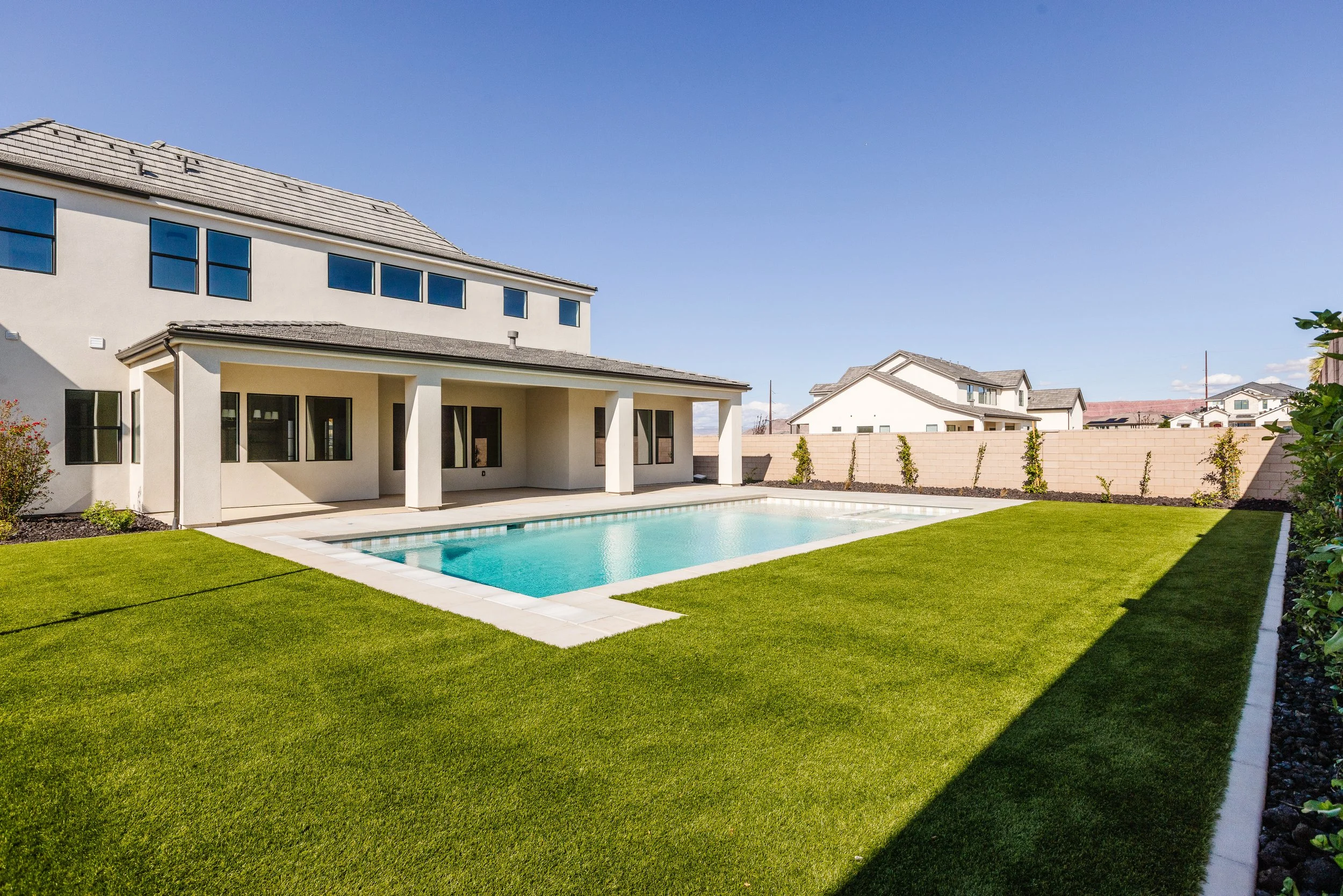 A modern house with a backyard swimming pool, surrounded by a green lawn and small plants along the fence, under a clear blue sky.