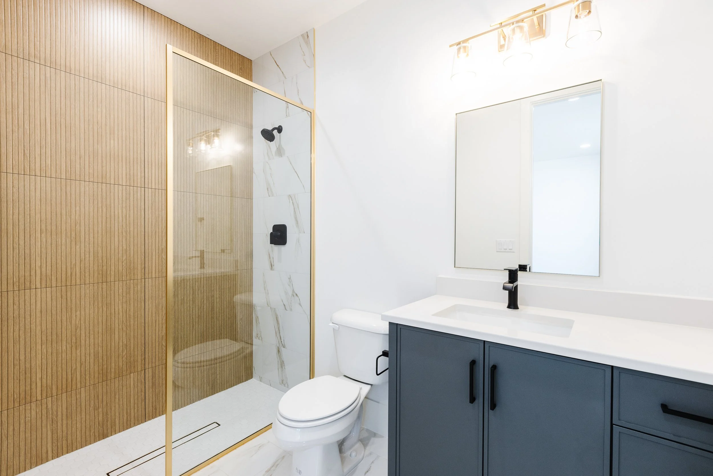 Modern bathroom with a walk-in shower with wooden and marble tiles, a white toilet, a blue vanity with a white countertop, a mirror, and a window.