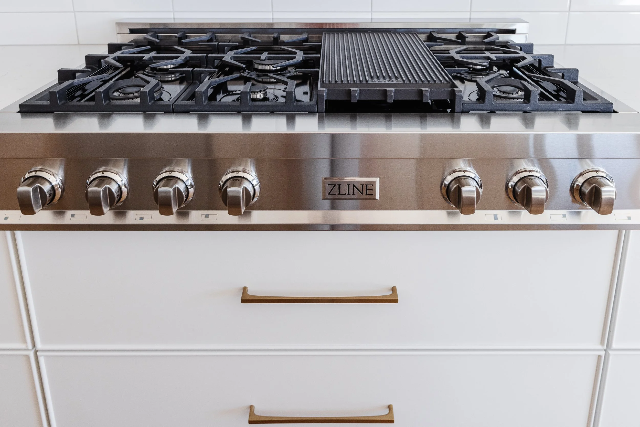 A modern stainless steel gas stove with six burners and a griddle in the center, set against a white tiled wall.