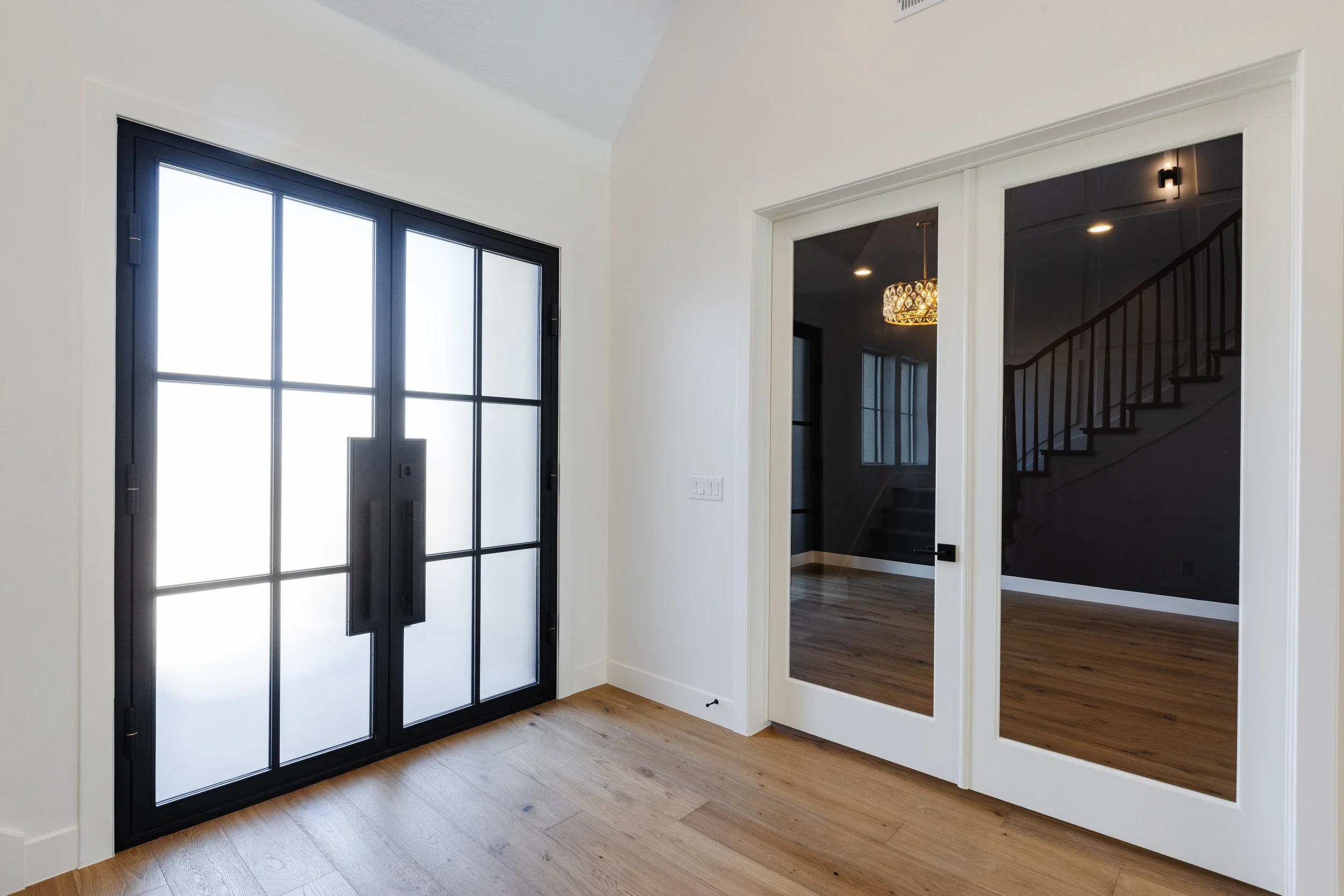 Entryway with black-framed glass door and adjacent wall-mounted mirror showing staircase and chandelier inside modern home.