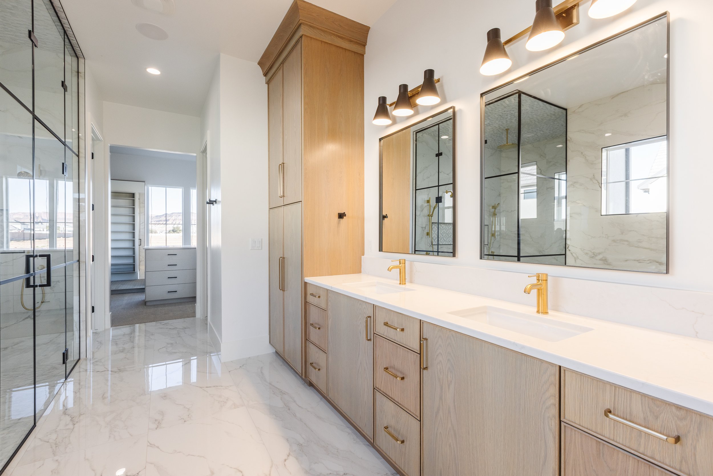 Modern bathroom with a large mirror, wooden cabinets with gold handles, and a white marble countertop with gold faucets. There are two separate shower areas with glass doors, one with a black grid pattern and the other with a marble wall. The floor a