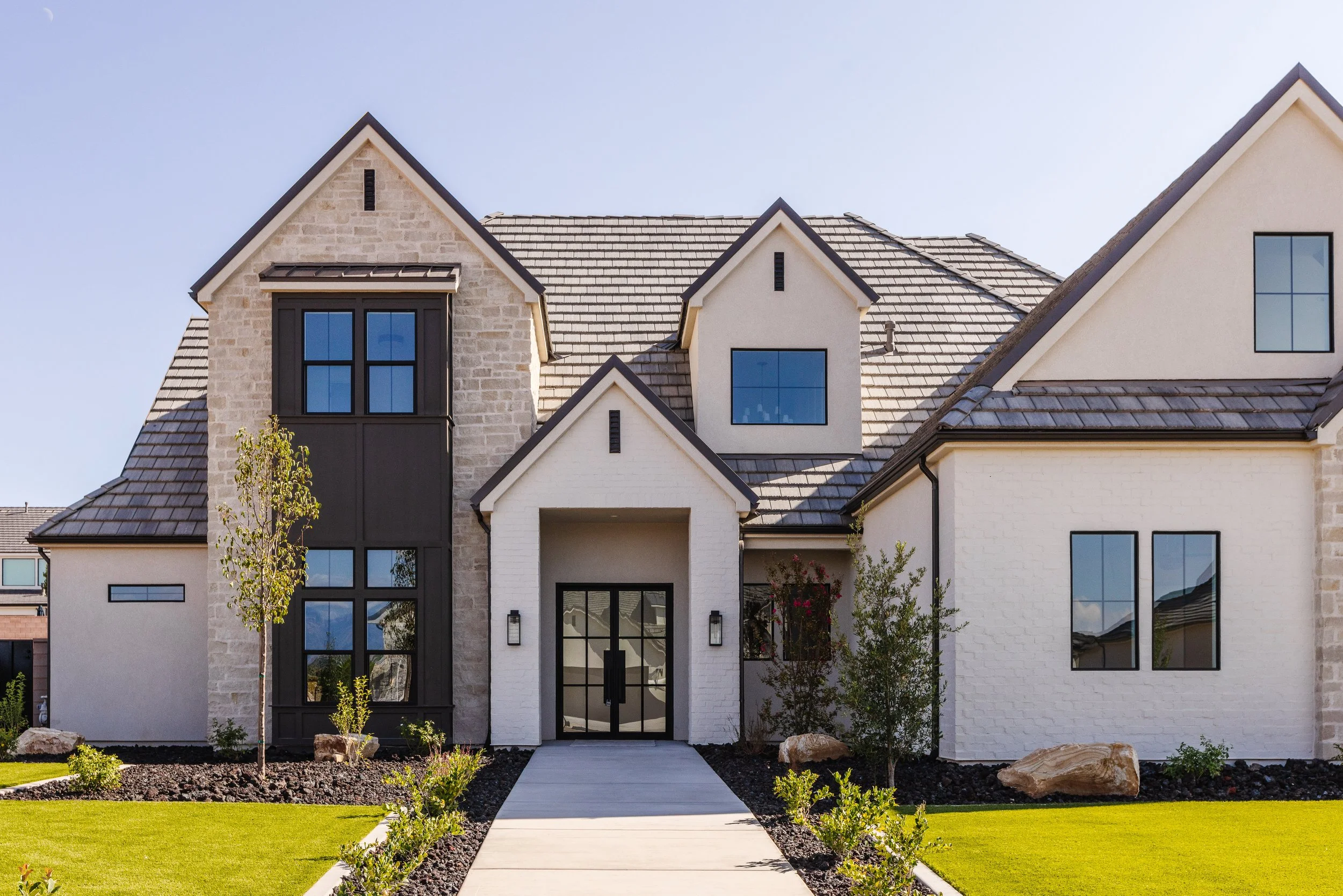 Front view of a modern house with a combination of brick and stucco exterior, steep gable roofs, large windows, and a landscaped front yard with a sidewalk.