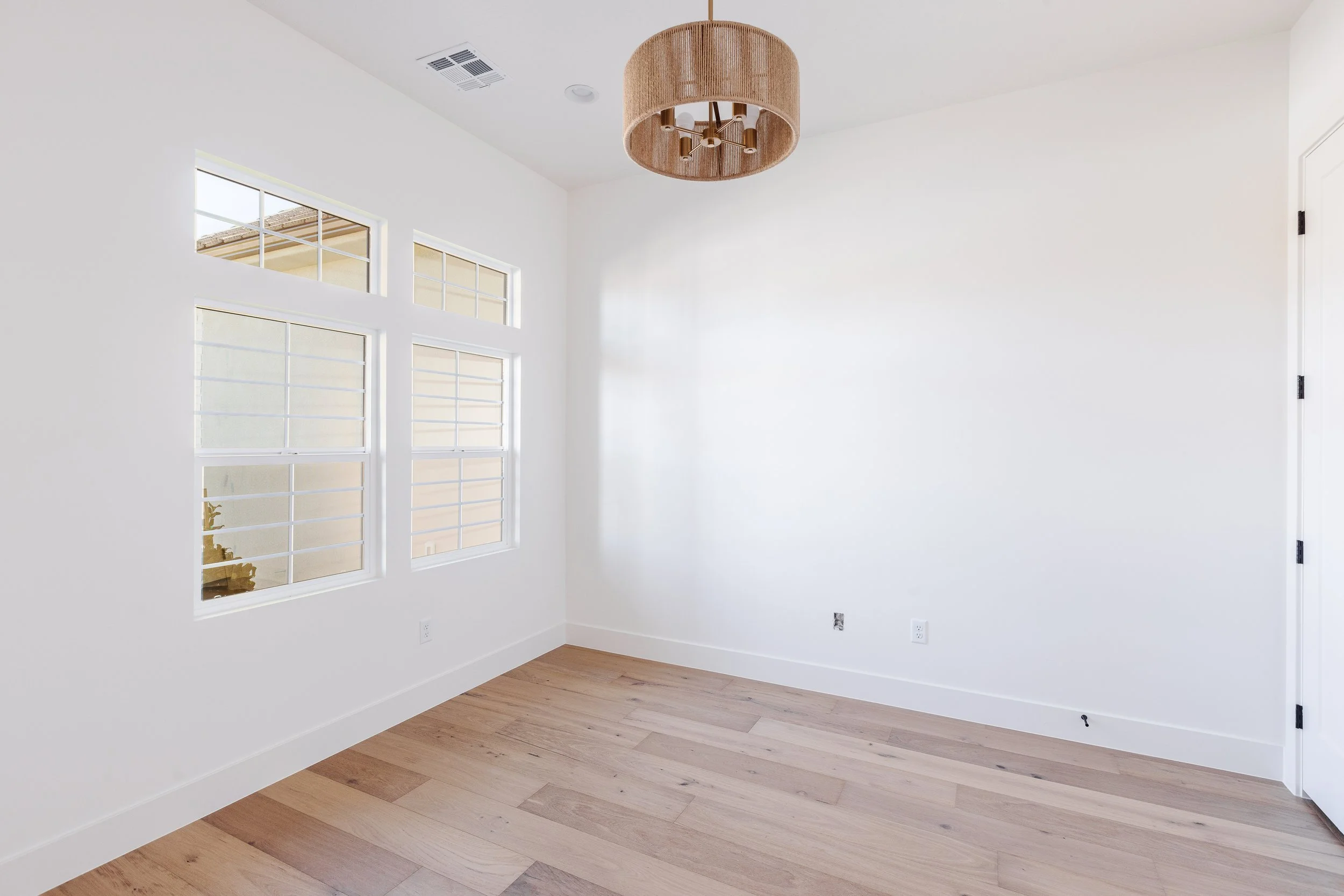 Empty room with white walls, hardwood flooring, multiple large windows, a ceiling light fixture, and a closed door.