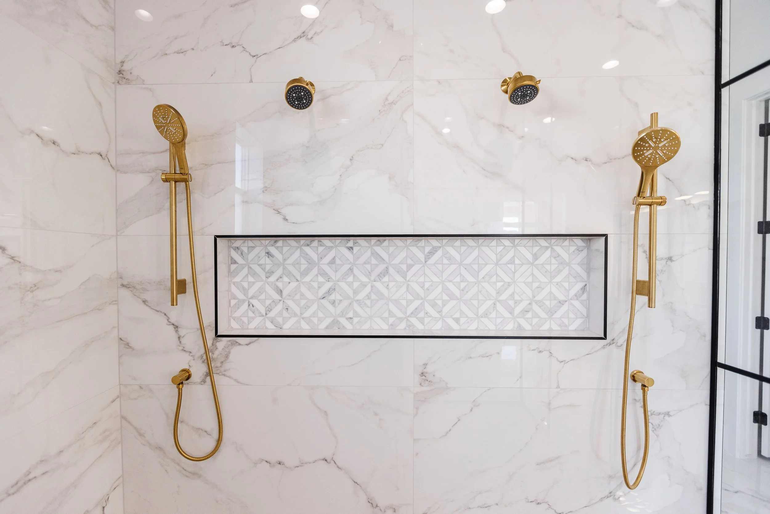 A modern shower with two gold shower heads and handheld sprayers mounted on a white marble wall with gray veins. There is a built-in niche with geometric tile pattern in the wall.