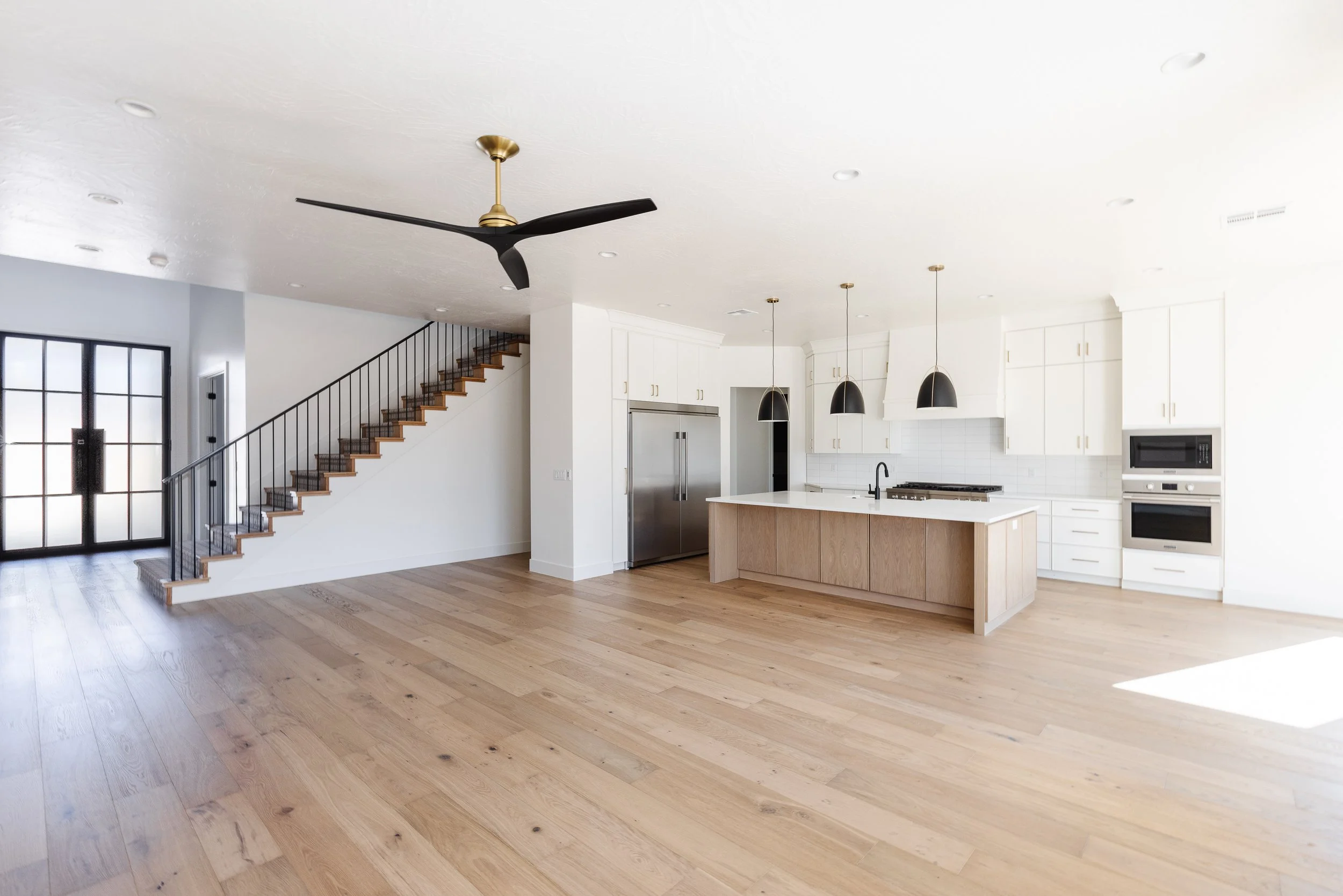 Open-concept kitchen and living area with light wooden floors, white cabinets, black pendant lights, a black ceiling fan, and a staircase with black railing leading upstairs.