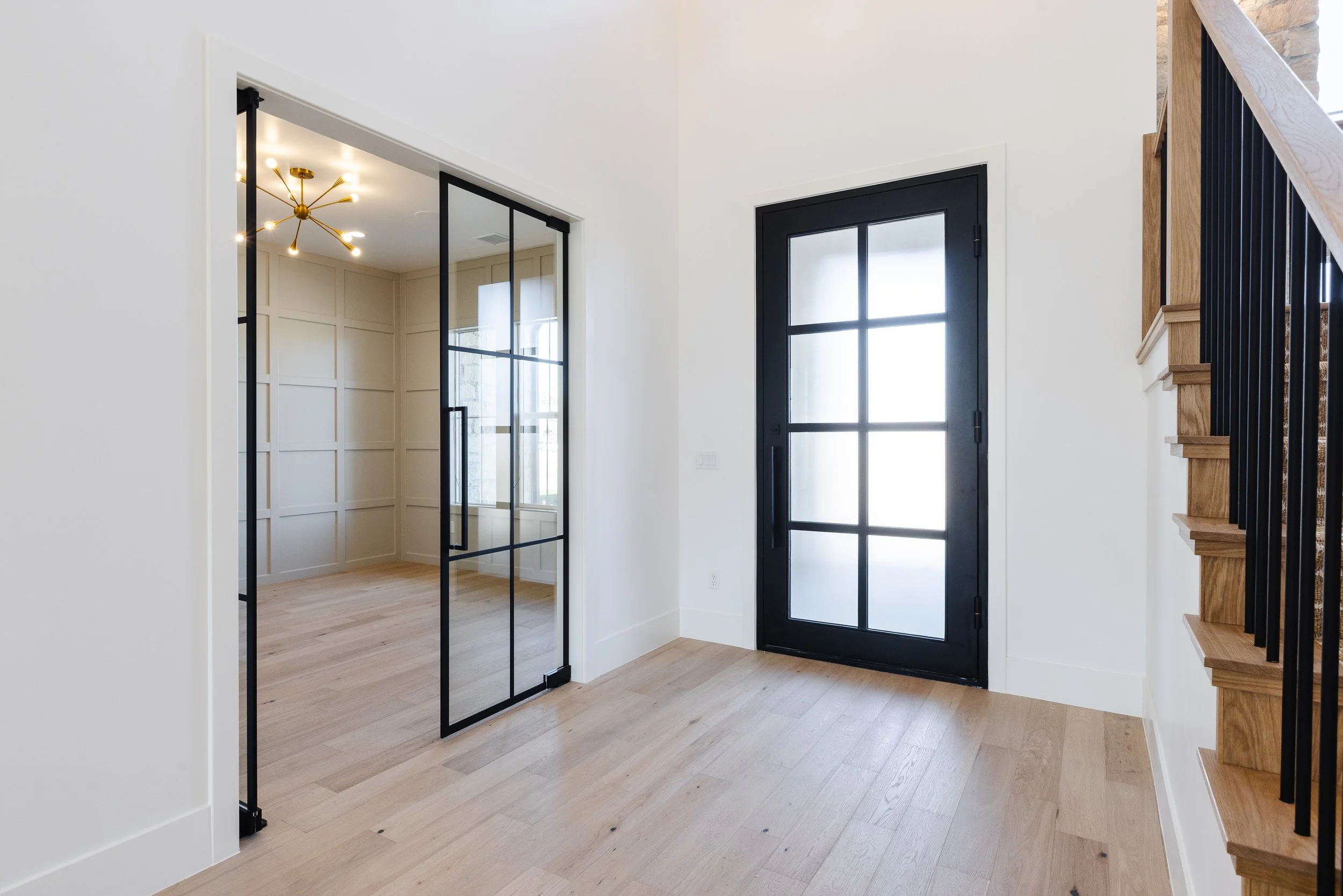 Interior view of a modern house entryway with a black framed glass door, a staircase with wooden steps and black metal railing, a glass sliding door leading to another room, and light wood flooring.
