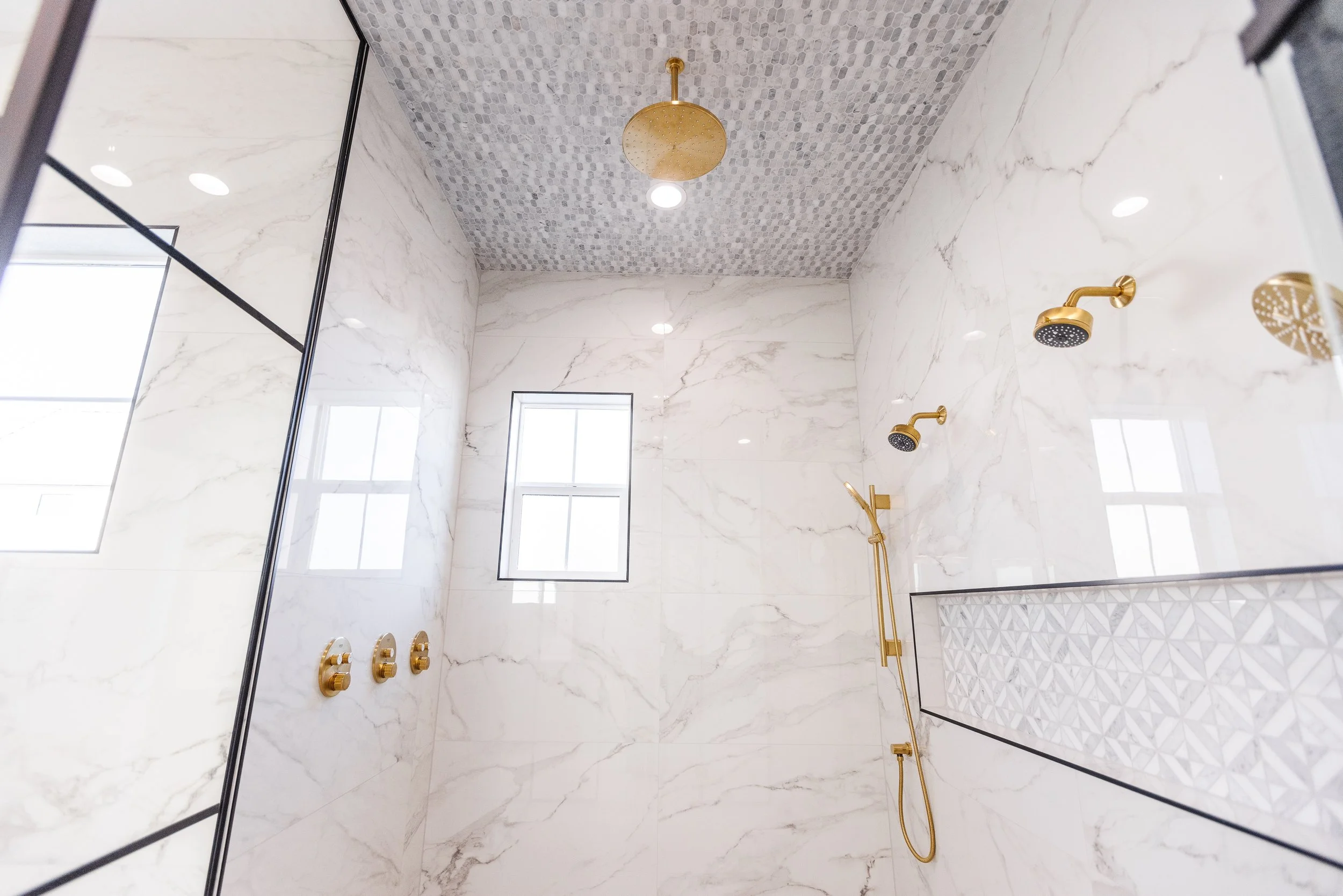 A modern walk-in shower with marble walls, a decorative tile ceiling, and gold fixtures including multiple showerheads and controls. There are two small windows providing natural light.