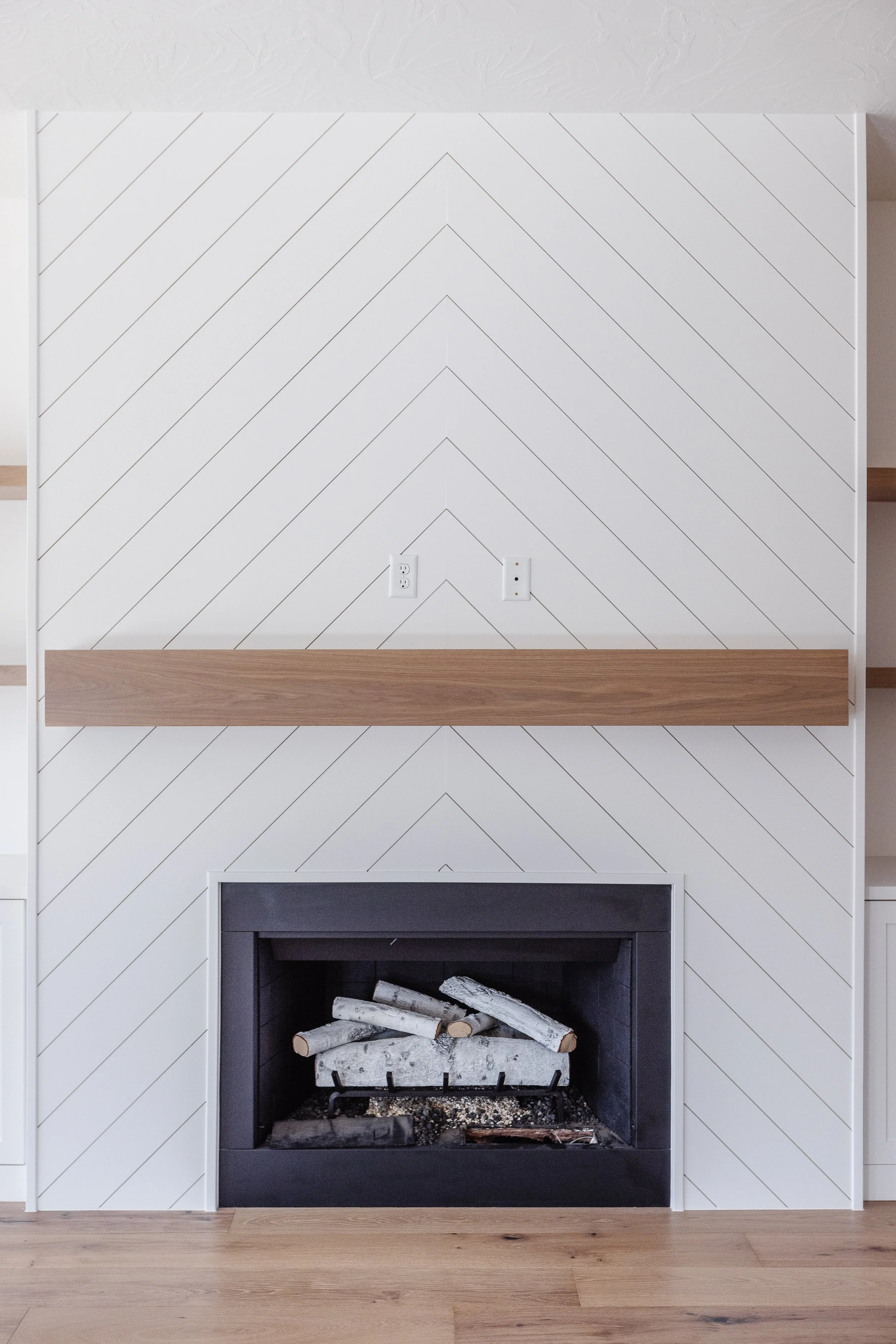 Interior of a living room fireplace with a modern black surround, white chevron-patterned wood paneling wall, a wooden mantel shelf, and a pile of logs inside the fireplace. Light wooden floor.
