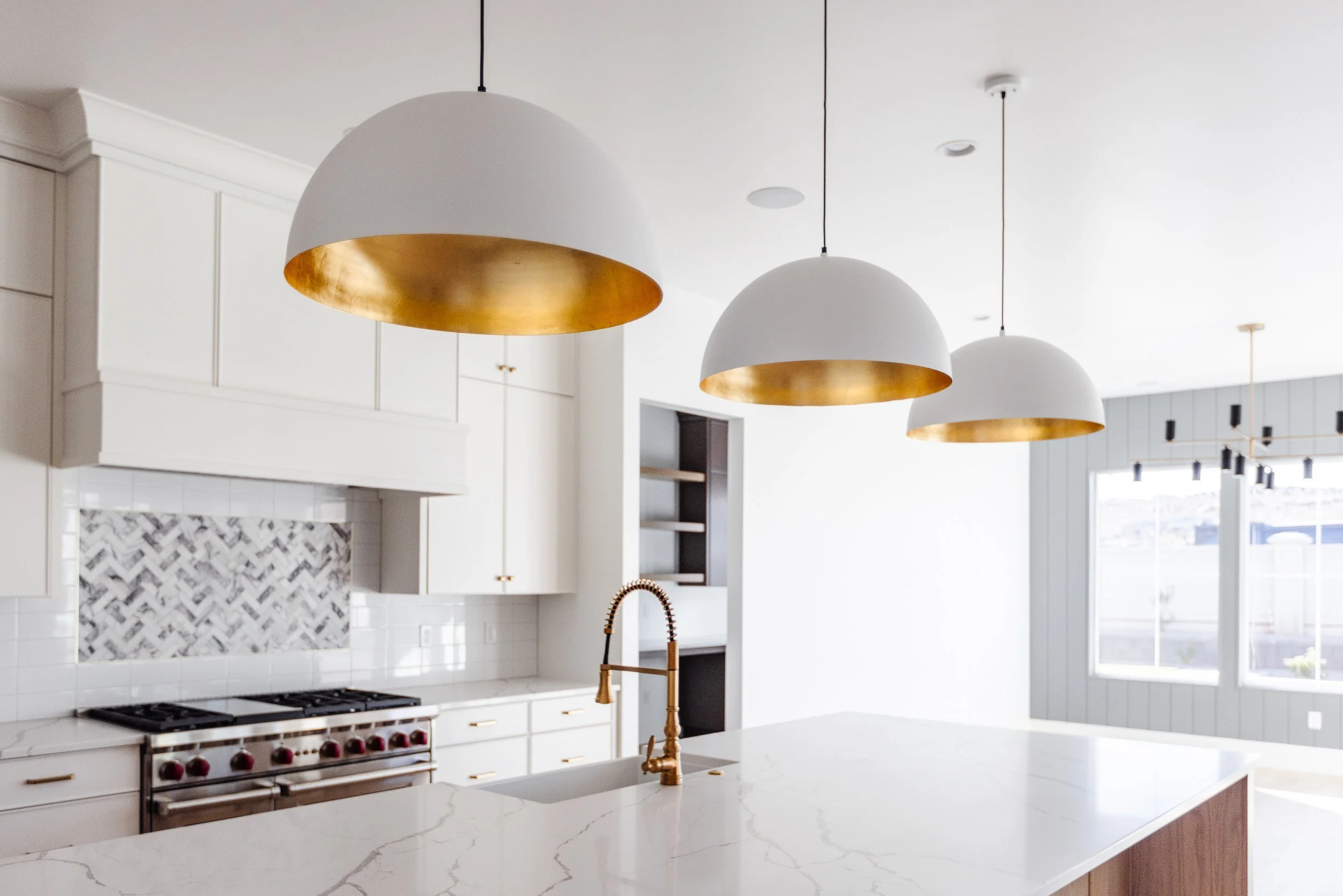 Modern kitchen with white cabinets, marble island counter, gold sink faucet, black and gold stove, pendant lights with white and gold interiors, and a window with natural light.