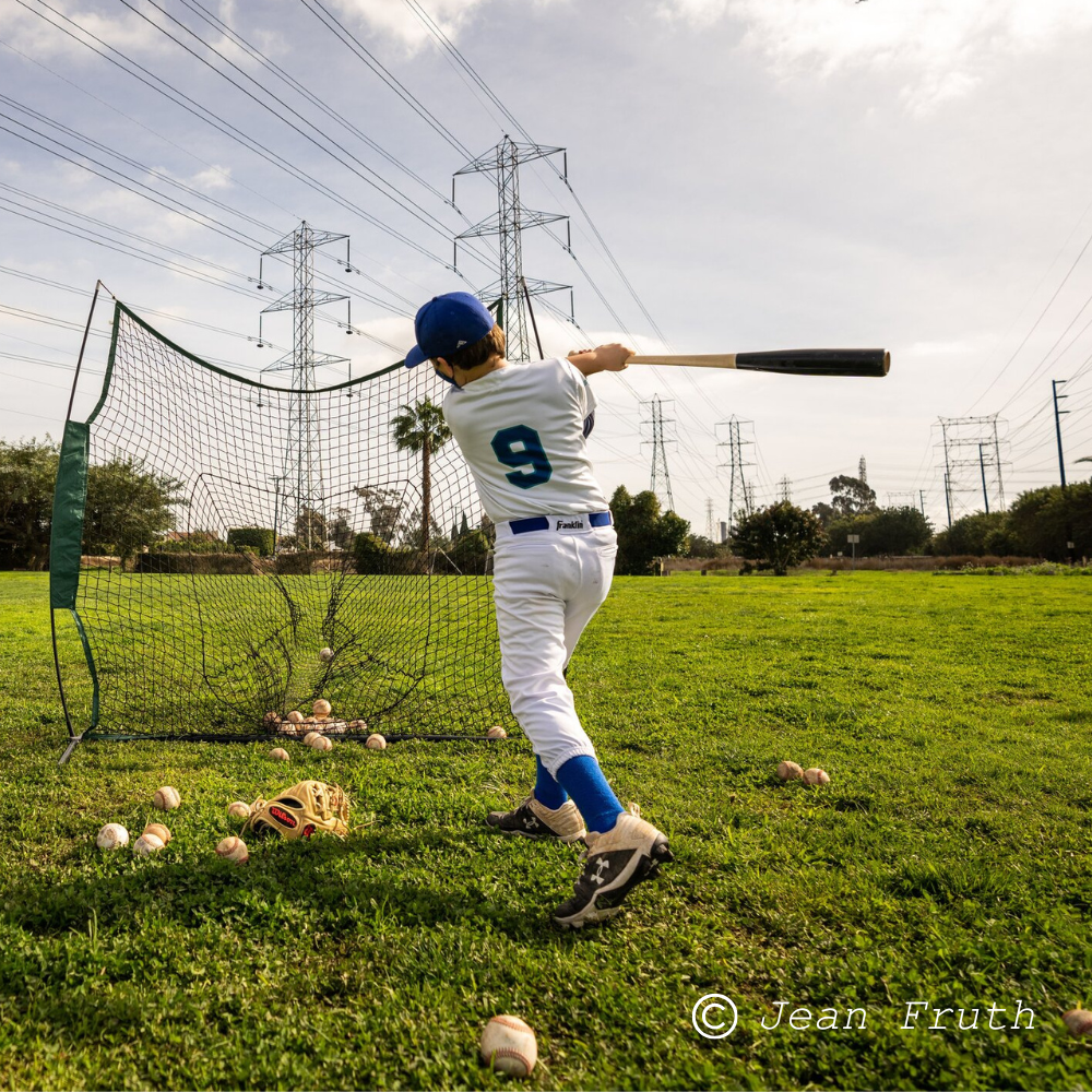 An athletic boy in baseball uniform, wearing a blue cap and jersey, practicing batting in a park. He is swinging a baseball bat toward a pitching screen surrounded by scattered baseballs on the grass under a cloudy sky.