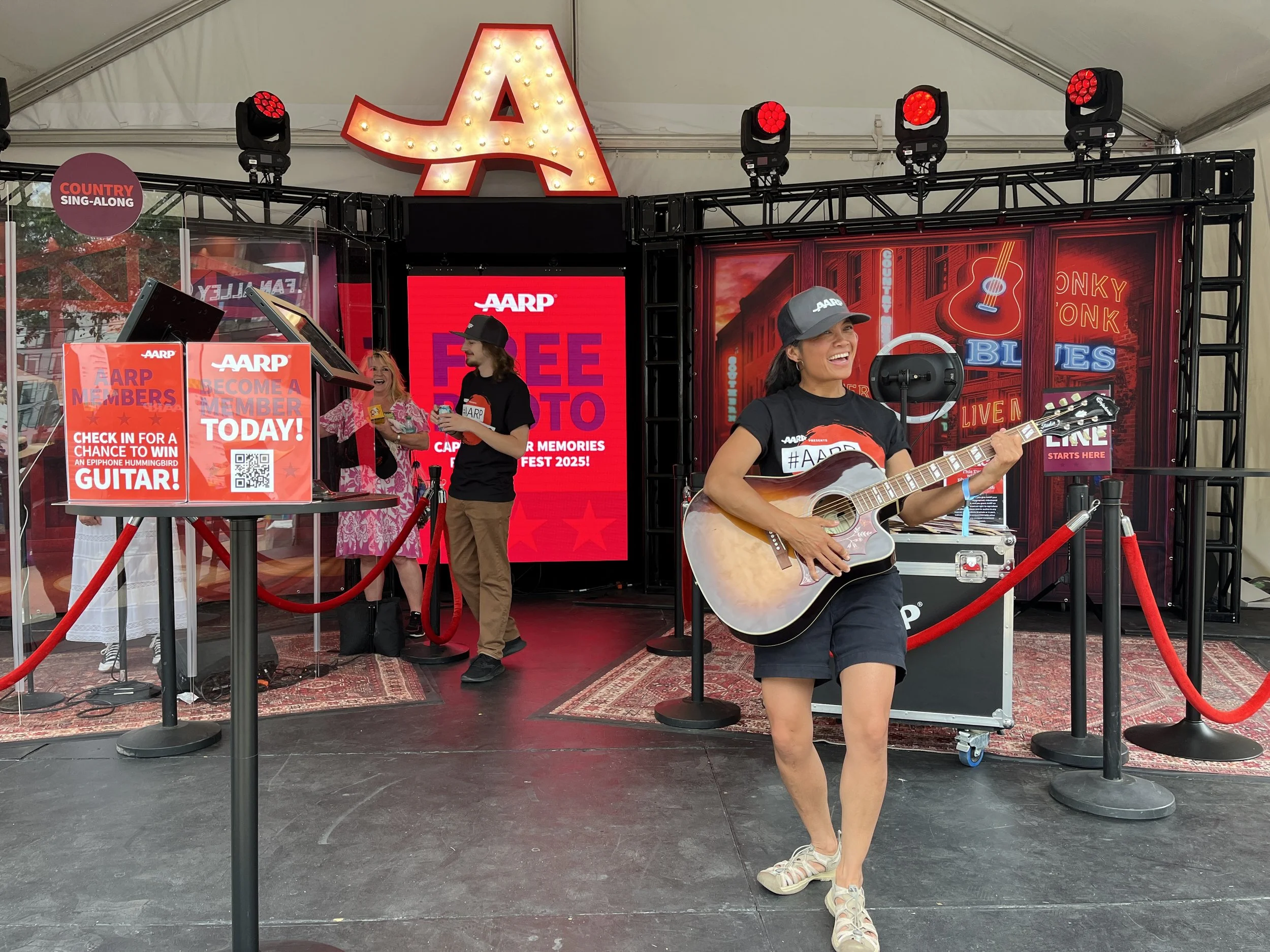 AARP booth at CMA Fest 2025 featuring a woman playing guitar with a photo booth behind her. The stage features red lights and branding and AARP signup information