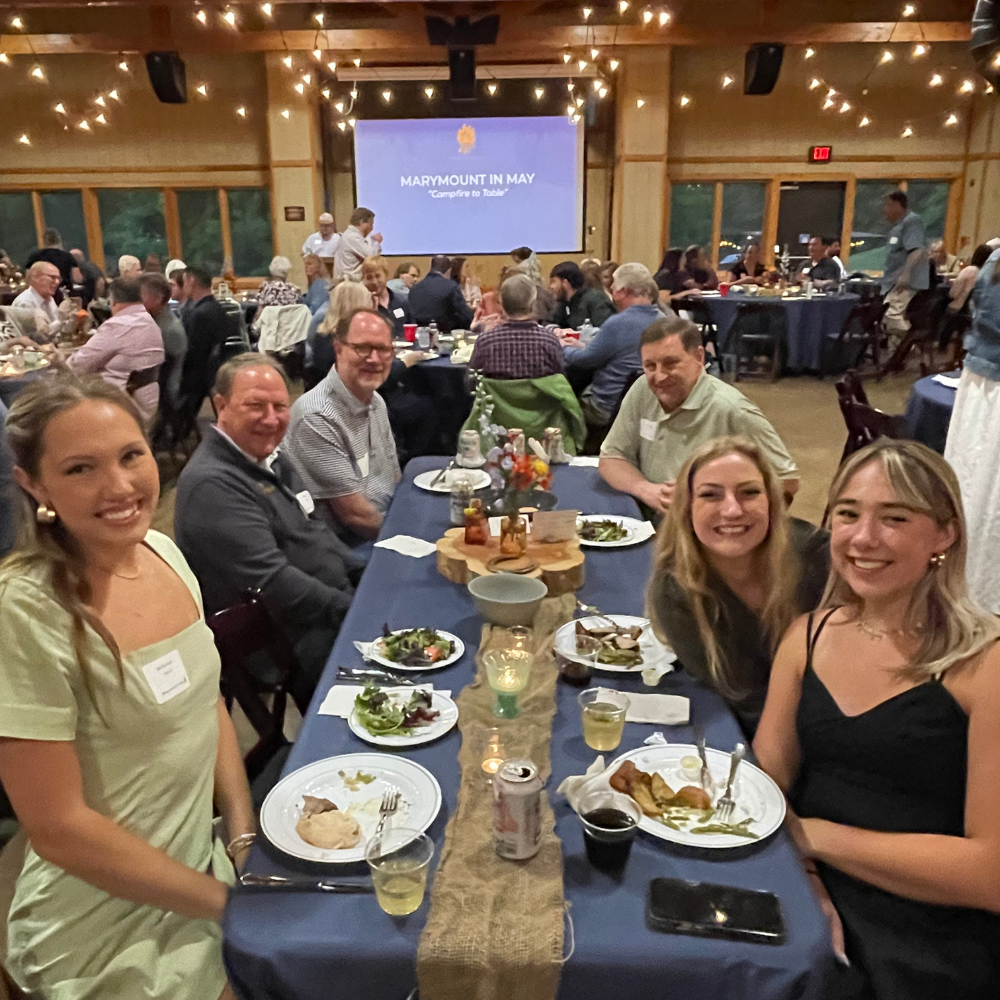 A smiling group at a nonprofit fundraiser seated around a long table with plates of food, drinks, and a centerpiece. In the background, a projector screen displays the text 'Marymount in May' for Camp Marymount's gala and auction outside Nashville.