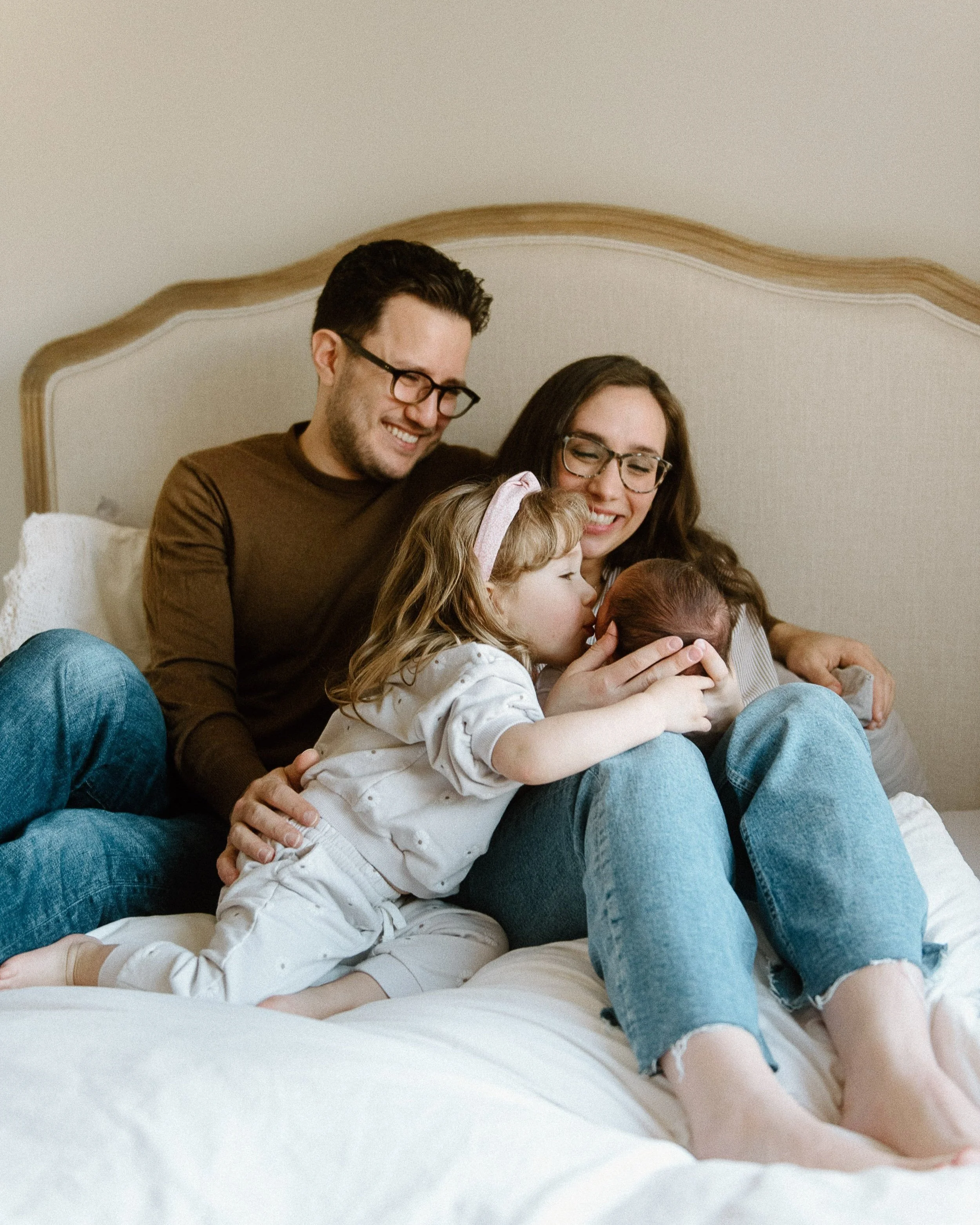 Young daughter kissing newborn while parents smile on bed.