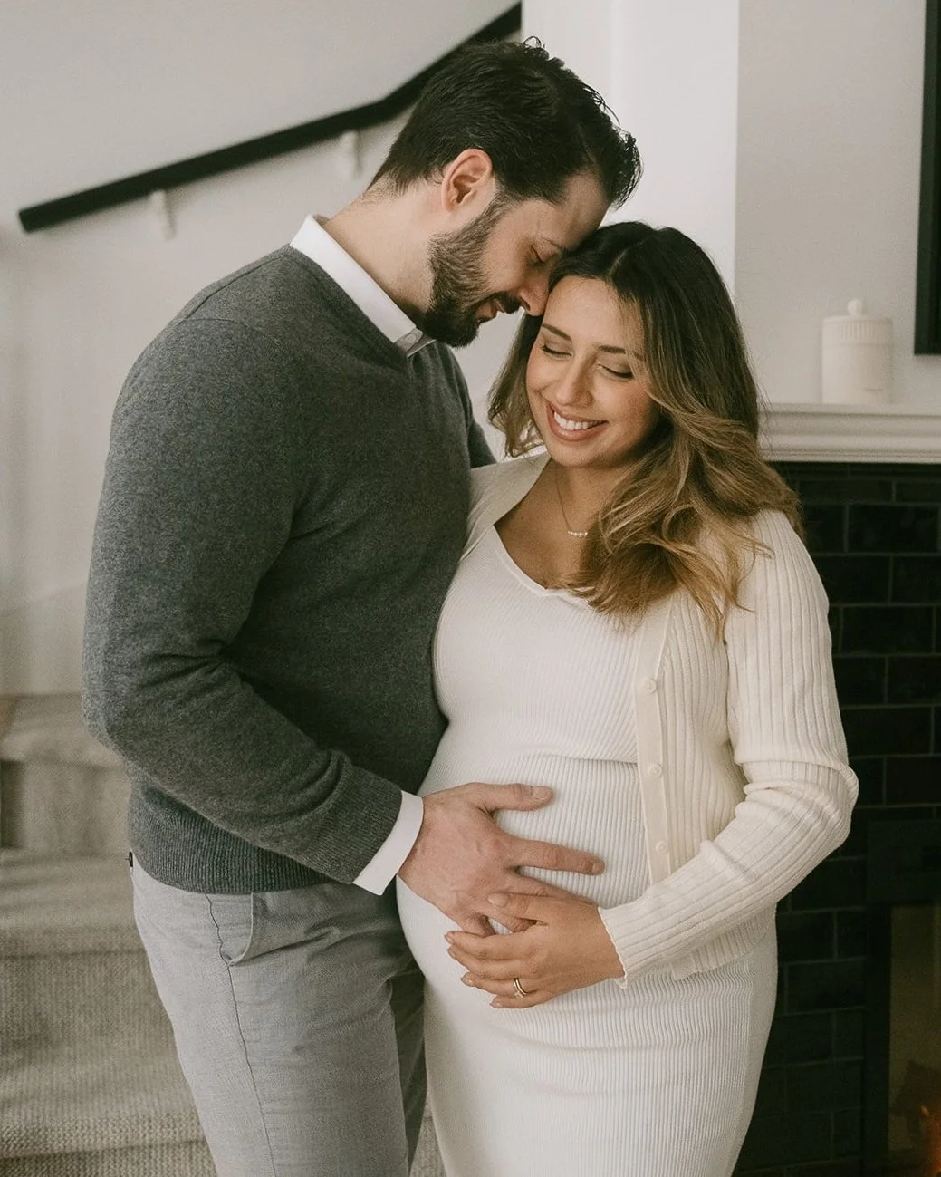 Husband resting head on pregnant wife's head next to fireplace.