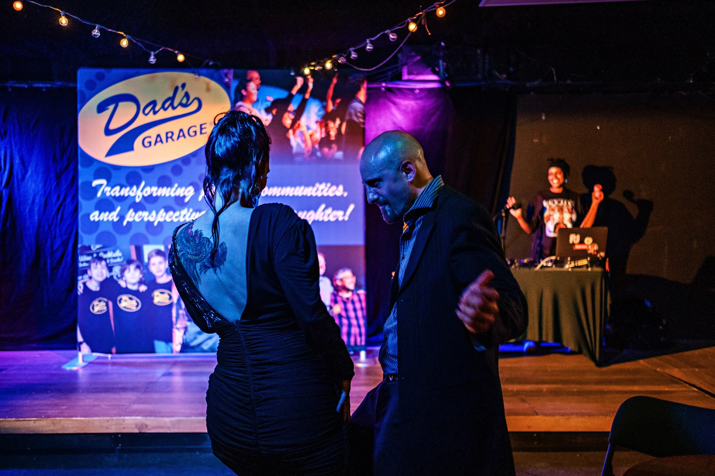Two people dancing together at an indoor event with a stage behind them, which displays a sign for Dad's Garage and a video of people dancing. Other people are visible on the stage, including a woman with a microphone and a DJ.