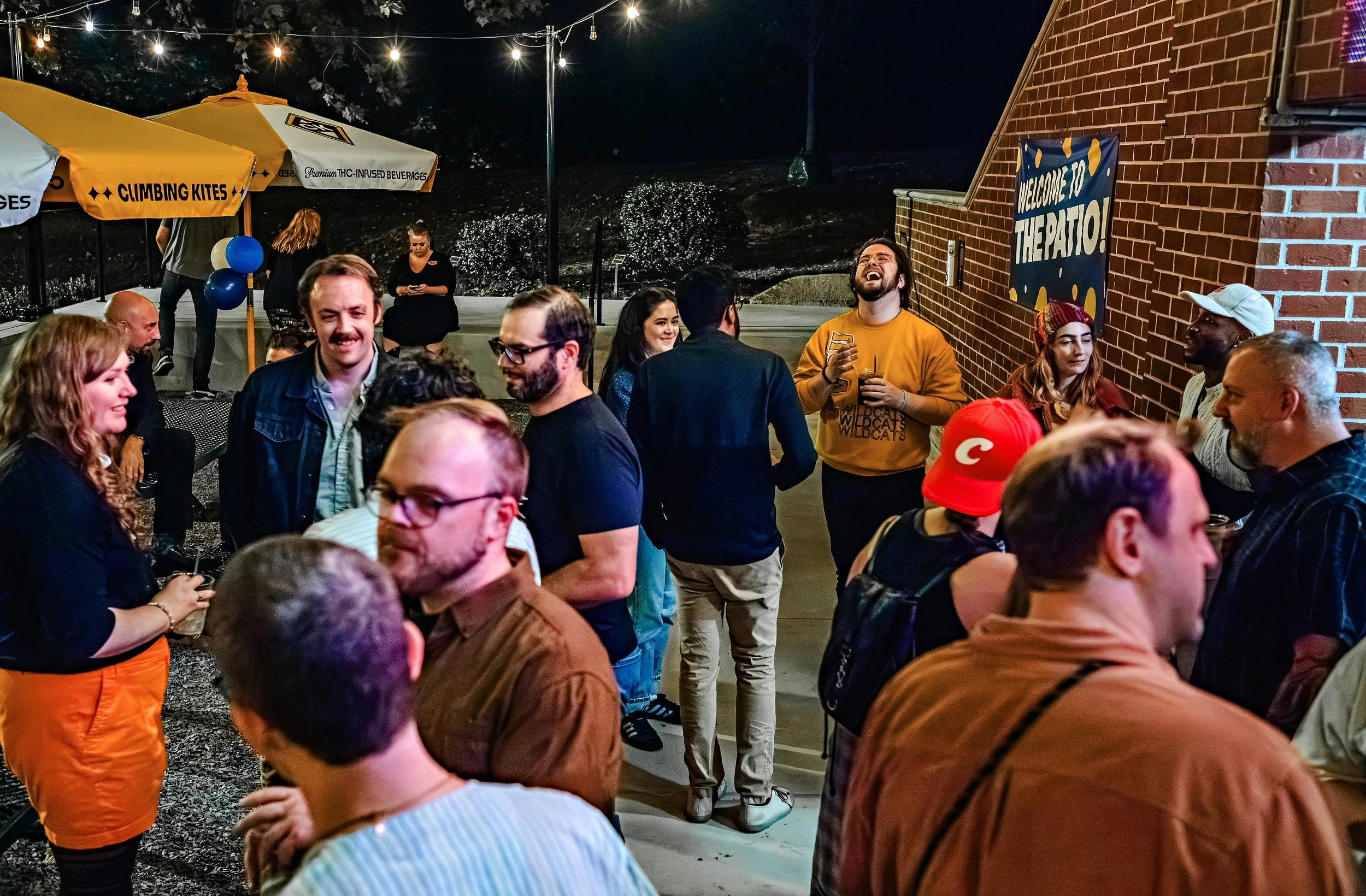 A group of people gathered at an outdoor event at night, with string lights overhead. Some people are chatting, while others are looking at their phones. There are yellow umbrellas with black text, balloons, and a brick building with a welcome sign that says 'Welcome to the Patio!'