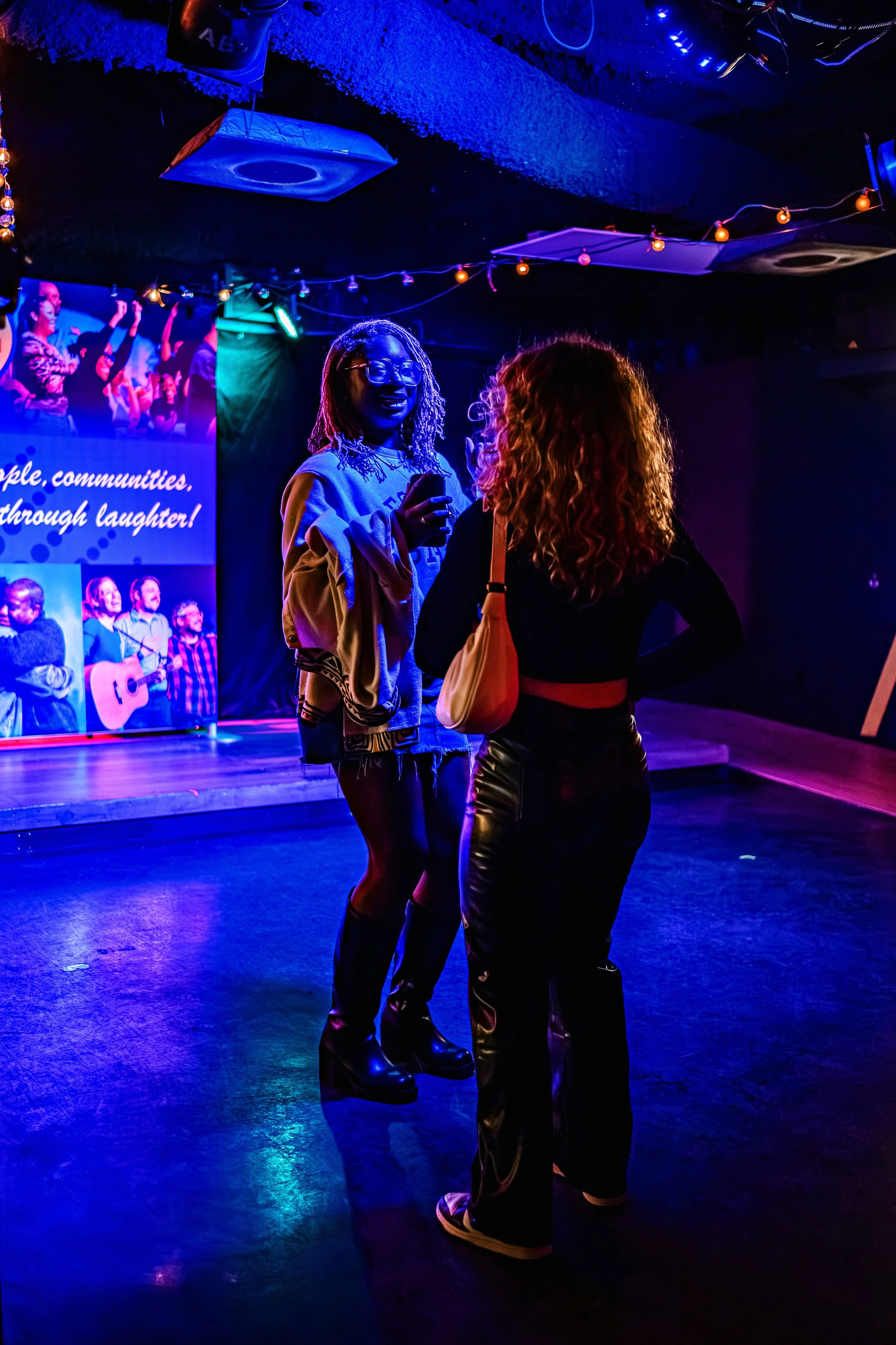 Two women standing and talking in a dimly lit room with blue and purple lighting, a stage with a musician playing guitar and singing in the background, and a large screen showing images of people dancing.