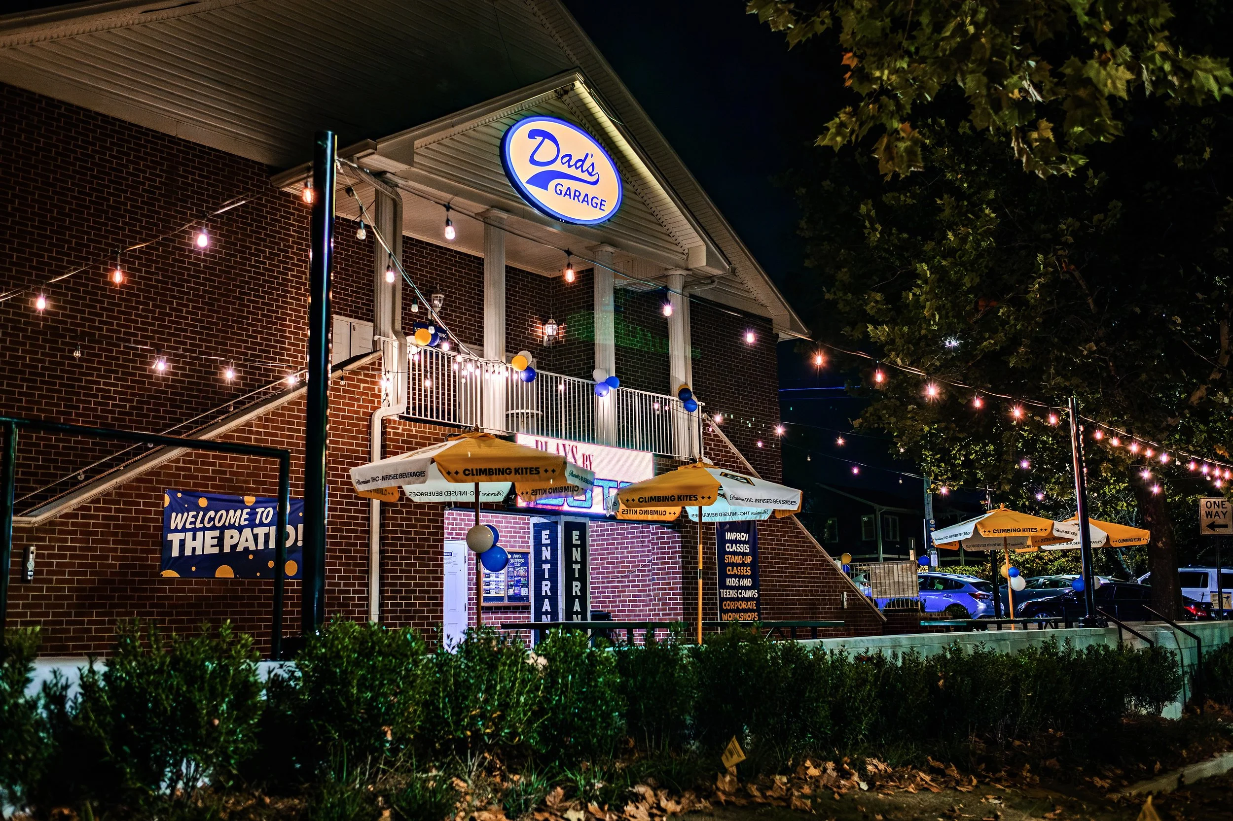 Nighttime exterior view of Dad's Garage building with illuminated sign, string lights, banners, and outdoor seating area decorated with balloons, surrounded by trees and parked cars.