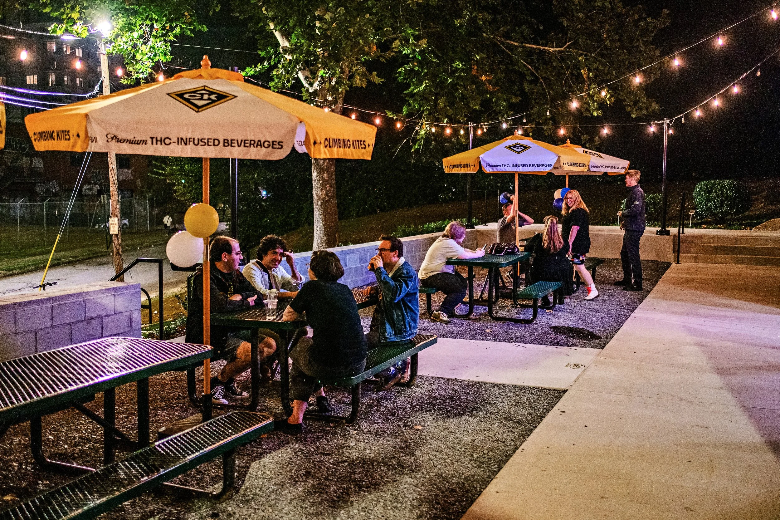 People sitting and standing at outdoor tables under umbrellas decorated with balloons, illuminated by string lights at night in a park.