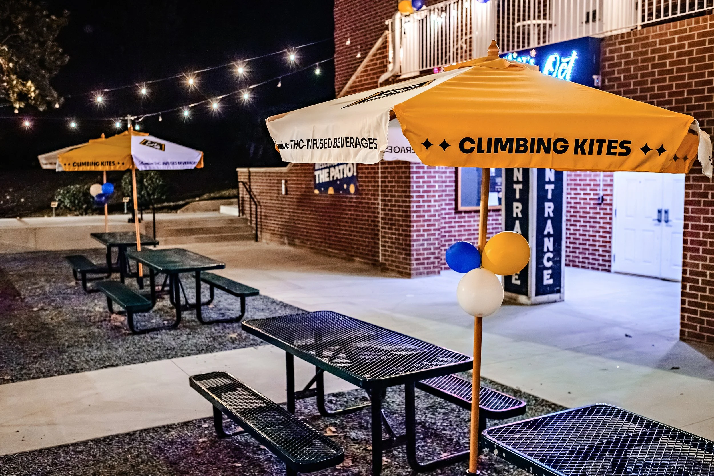 Nighttime outdoor patio area with black metal benches, yellow and white umbrellas with balloons, and string lights overhead. The umbrellas display text related to THC-infused beverages and climbing kites, with a brick building in the background and a neon sign partially visible.