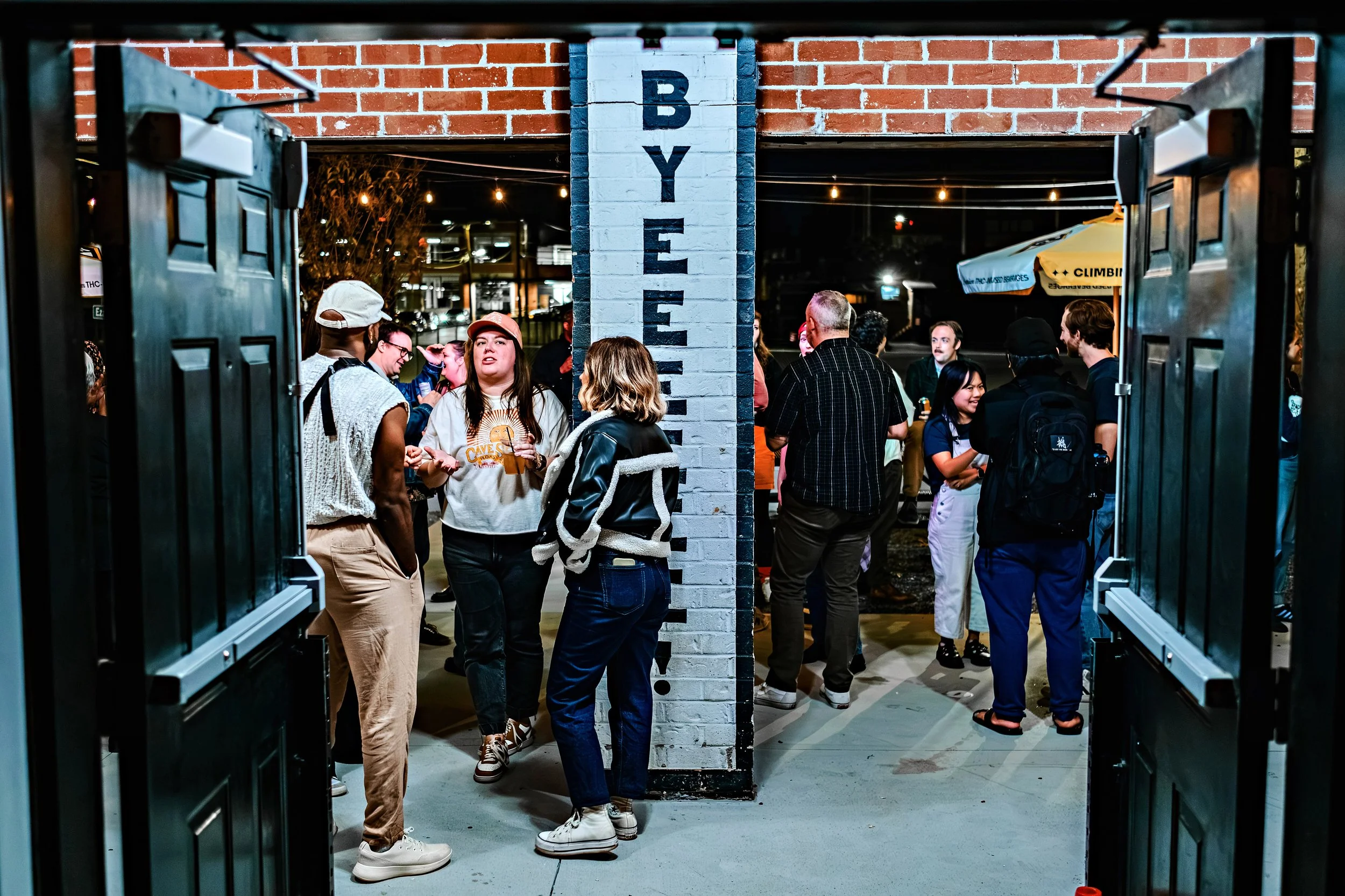 A group of diverse young people socializing at night inside a venue with a brick wall labeled 'BYE' and open doors, illuminated by warm string lights.