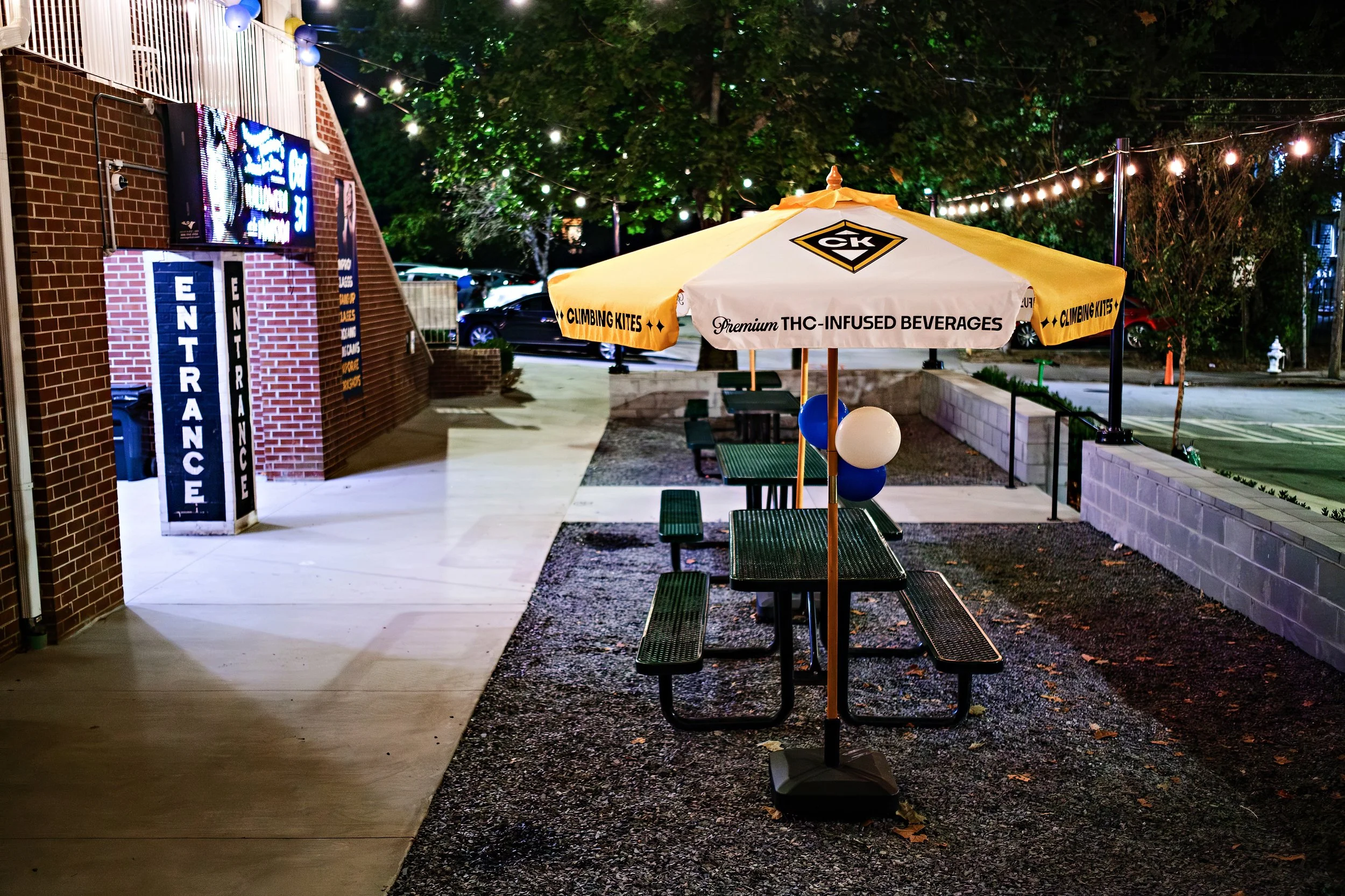 Nighttime outdoor scene with a large yellow and white umbrella advertising THC-infused beverages and climbing kites, surrounded by string lights, green trees, parked cars, and a brick building with a neon sign.