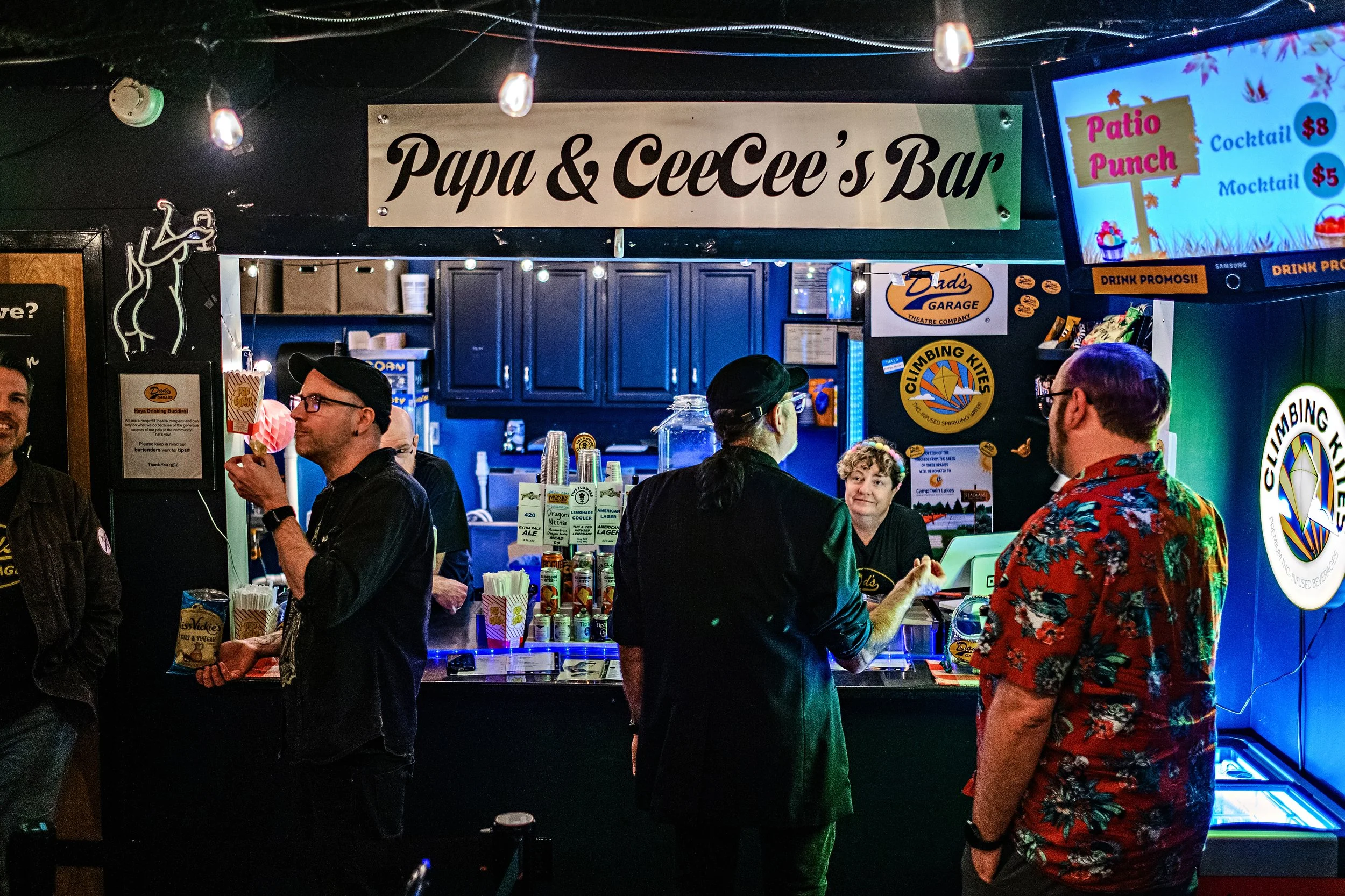 People standing at a bar called Papa & CeCe's Bar, with a sign above and a digital menu screen showing prices for cocktails and mojitos. The bar has a colorful and lively atmosphere.