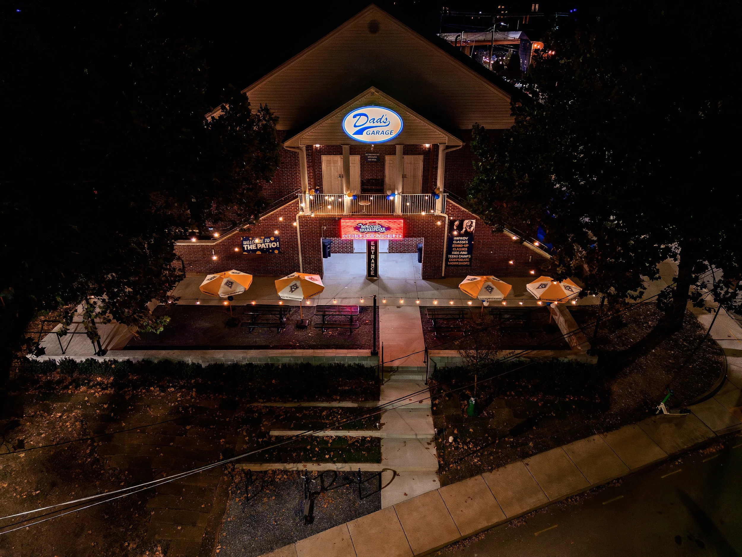 Night view of a brick building with a sign that reads Dad's Garage, decorated with string lights, and surrounded by outdoor tables and umbrellas.