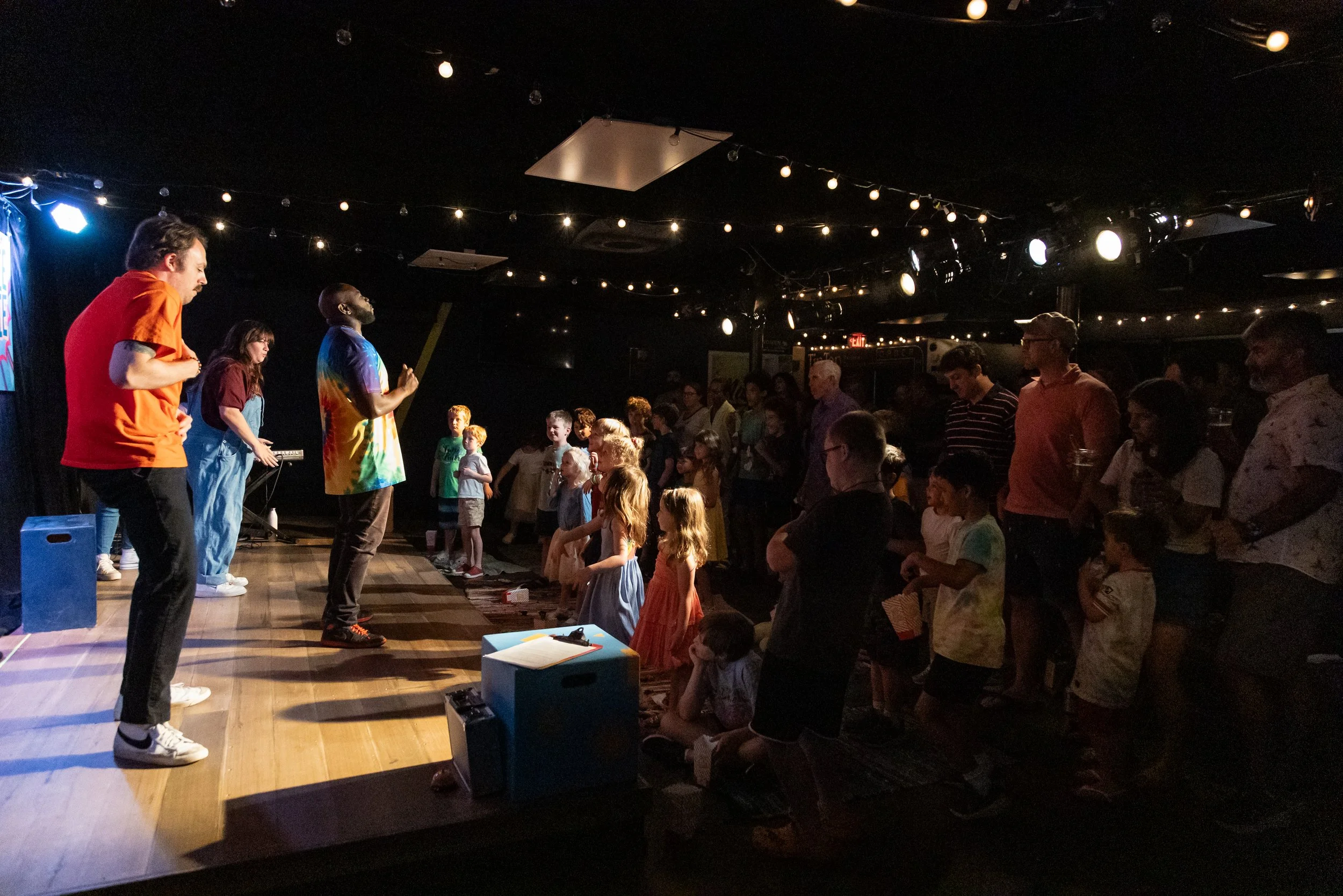 A live performance or event with a group of children and adults watching on a stage or in front of it, under stage lighting and string lights in a dimly lit room.