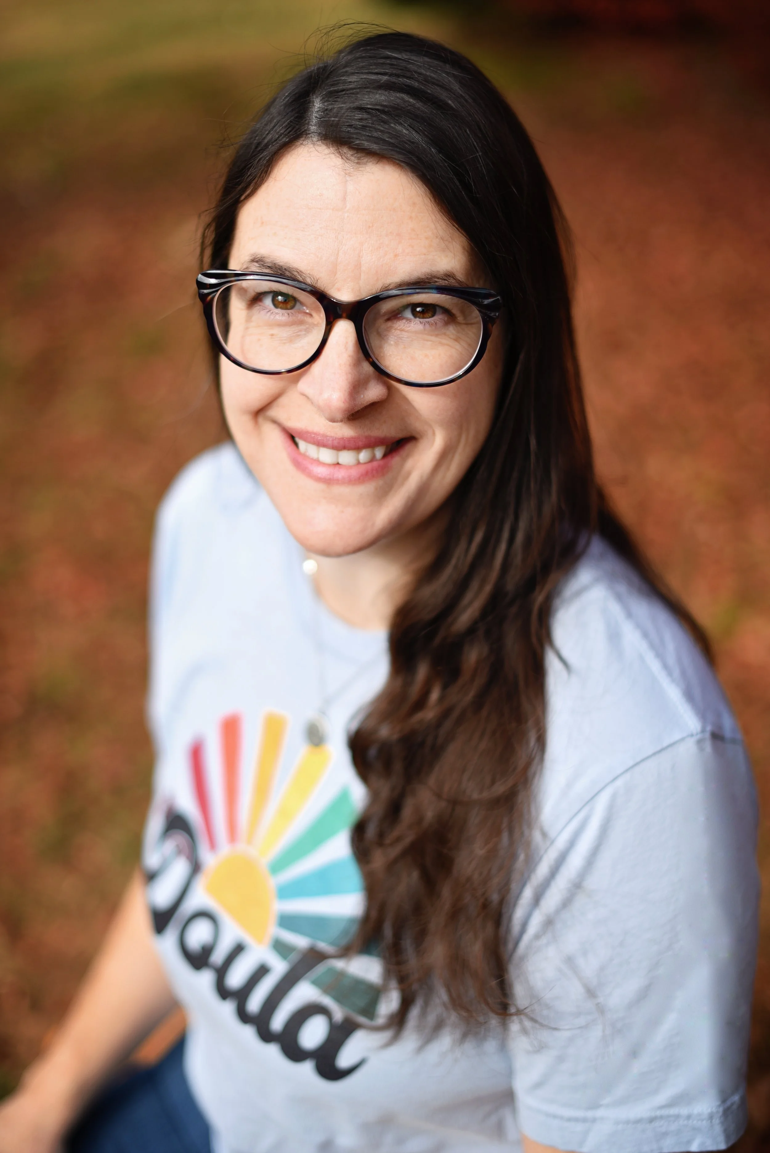 Abby Steffen, smiling with glasses and long brown hair, wearing a light-colored t-shirt with a colorful sunburst logo with the word "Doula", outdoors with a blurred autumn background.