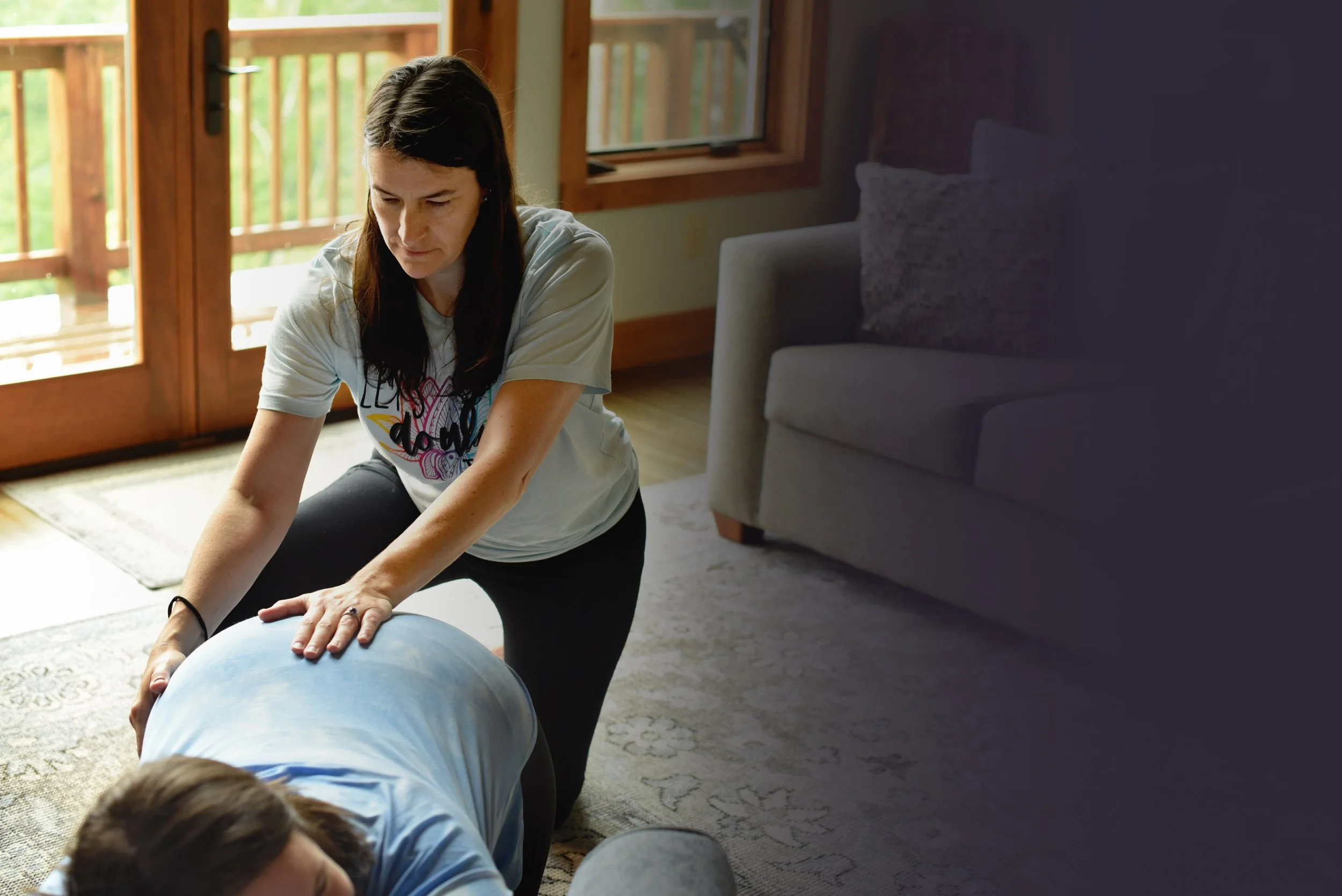 Abby Steffen is providing doula services to a woman in labor lying face down on the floor, with a large window behind them.