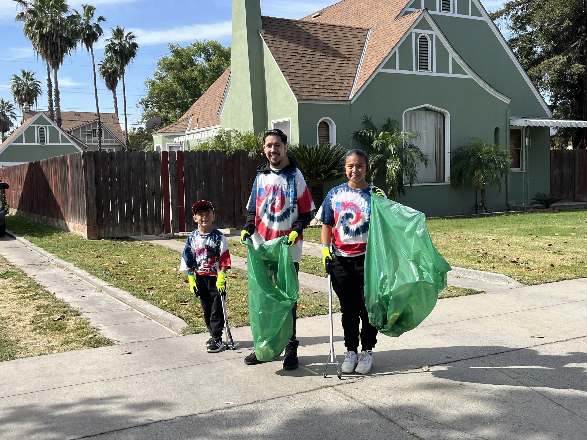 People participating in a community cleanup event, cleaning up debris and trash in an open lot near a fence, with some individuals using tools and wearing gloves, in an urban area with buildings, vehicles, and power lines in the background.