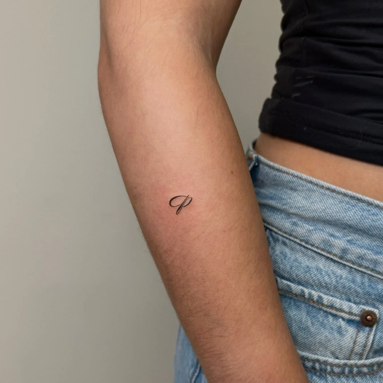 Close-up of a person's upper arm with a small, elegant tattoo of a cursive letter 'P' on the inner arm.