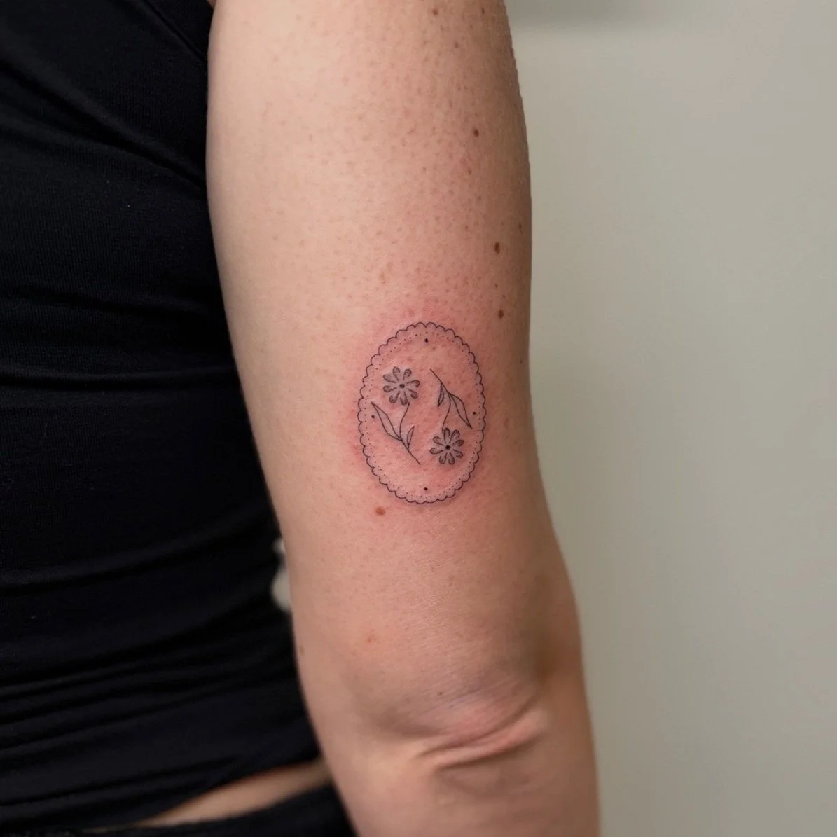Minimalist black ink tattoo of flowers and leaves inside an oval lace-like border on a person's upper arm.