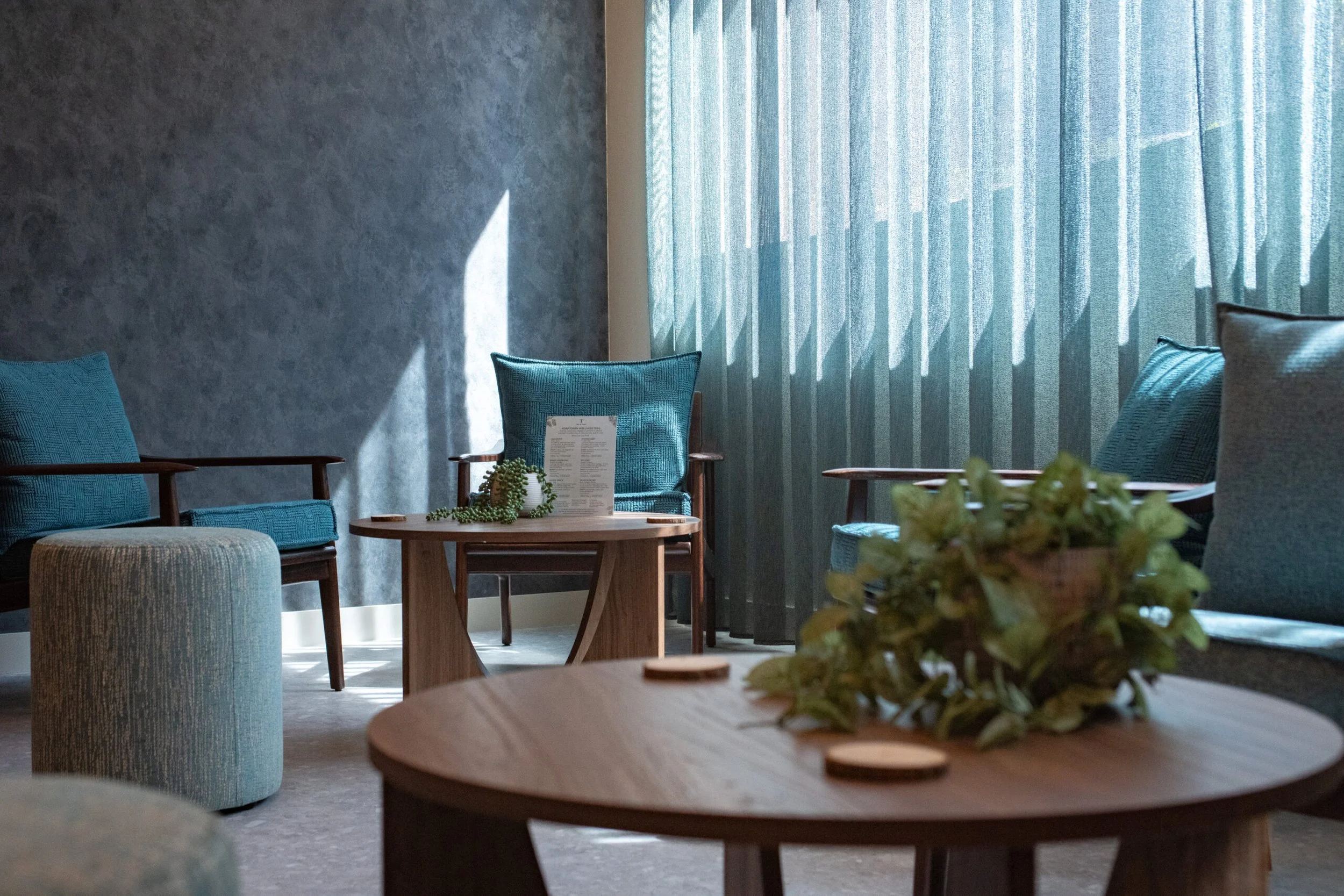 A welcoming relaxation lounge at Being Wellness Centre in St Albans, featuring comfortable seating, a wooden coffee table, natural greenery and soft daylight in a contemporary wellness setting.