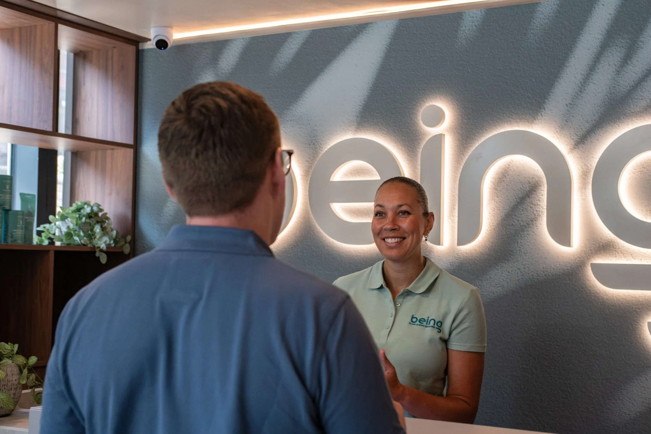 Alex James in conversation with a guest at Being Wellness Centre in St Albans, inside the contemporary reception area with the illuminated Being logo in the background.