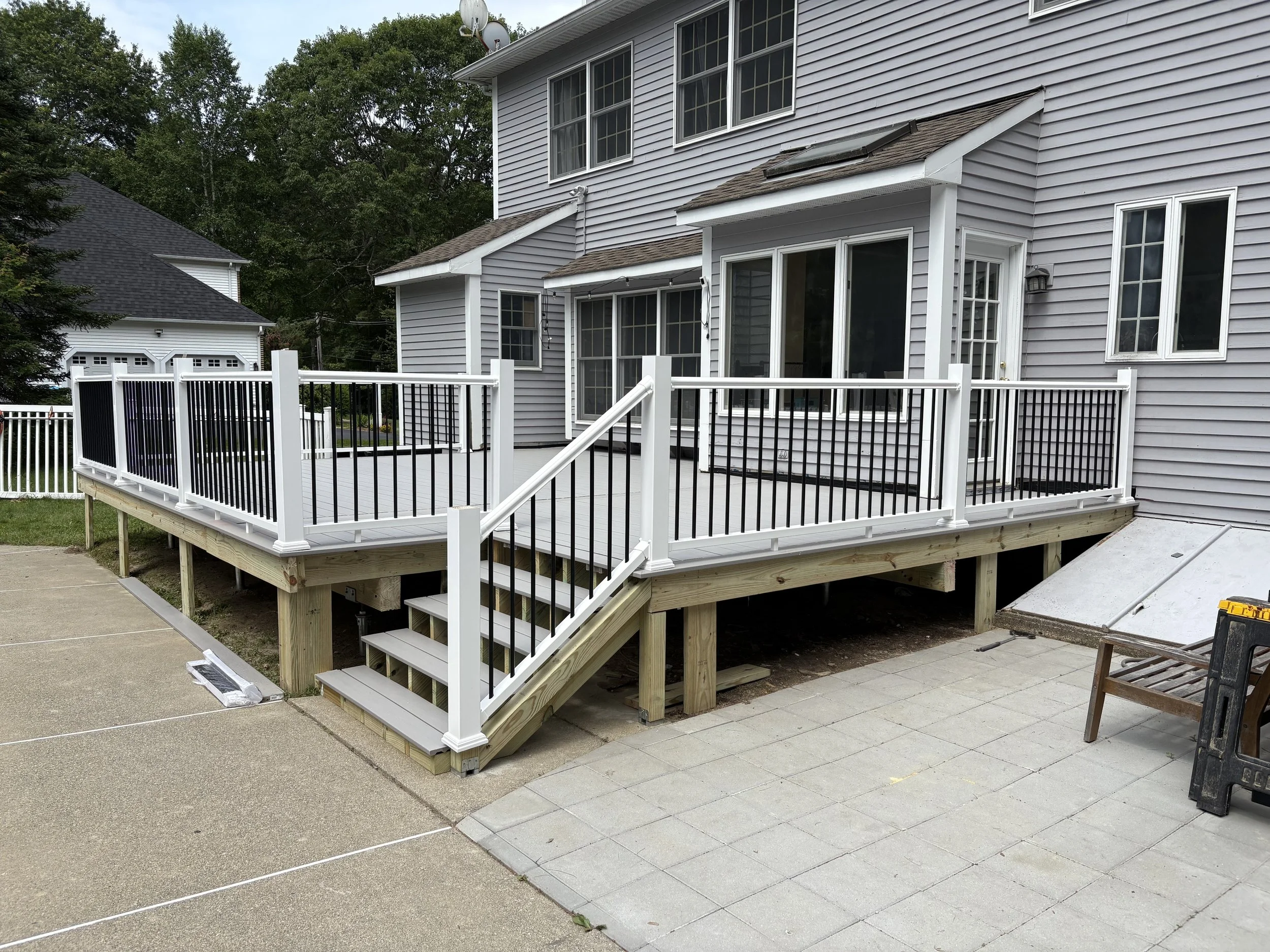 A newly built elevated wooden deck attached to a house with white and black railings, stairs leading down, and a sliding glass door, with concrete pavers underneath and a bench nearby.