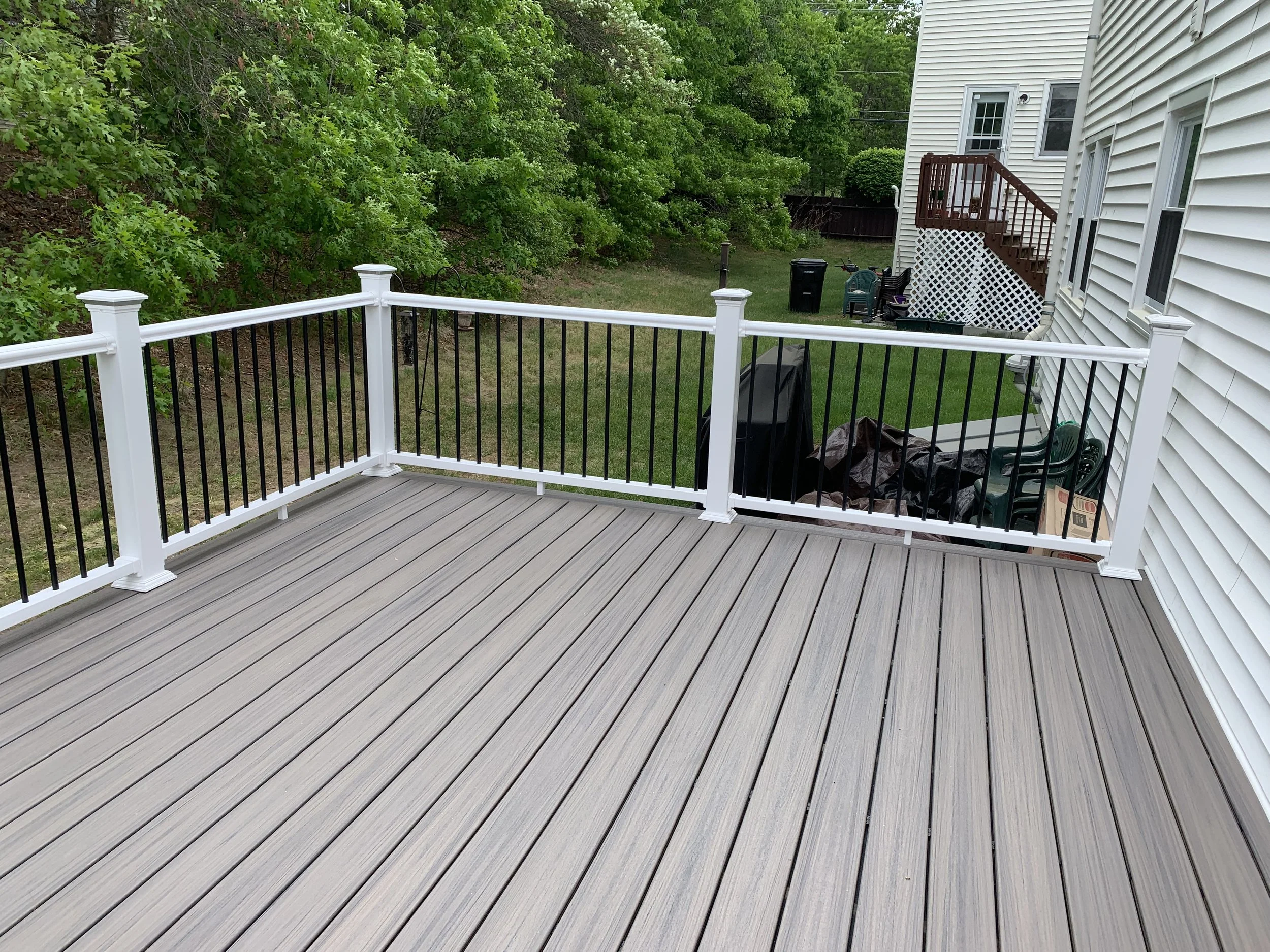 Backyard deck with white and black railing, beige wood flooring, trees on the left, and a white house with stairs and green lawn in the background.