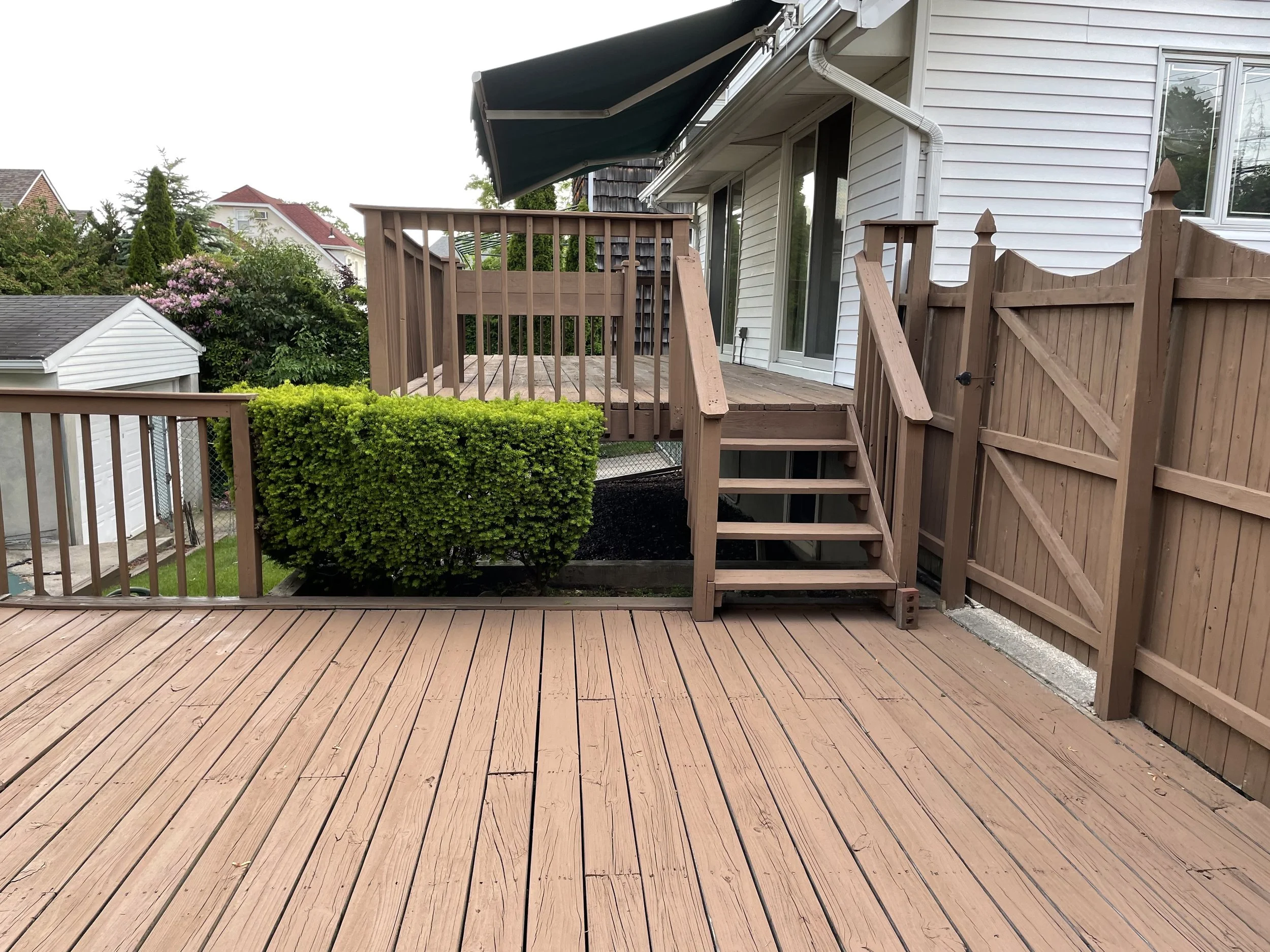 A backyard wooden deck with stairs leading to a lower level, surrounded by a wooden fence, with a white house with sliding glass doors and a green awning attached to the house. There is a small landscaped area with a bush and some mulch.