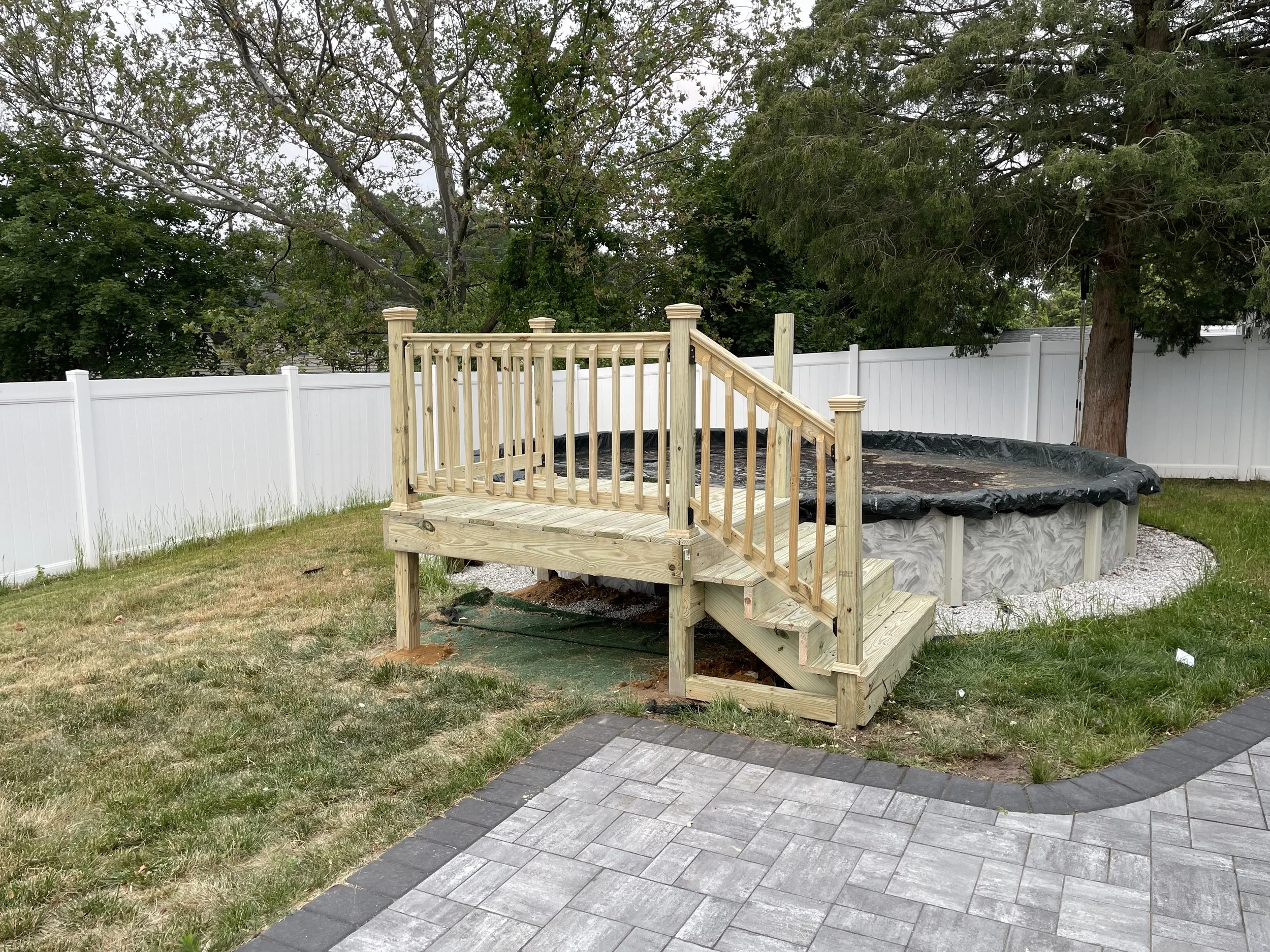 A backyard with an in-ground pool covered with black safety cover, a wooden deck with stairs and a railing, a white fence, and trees in the background.