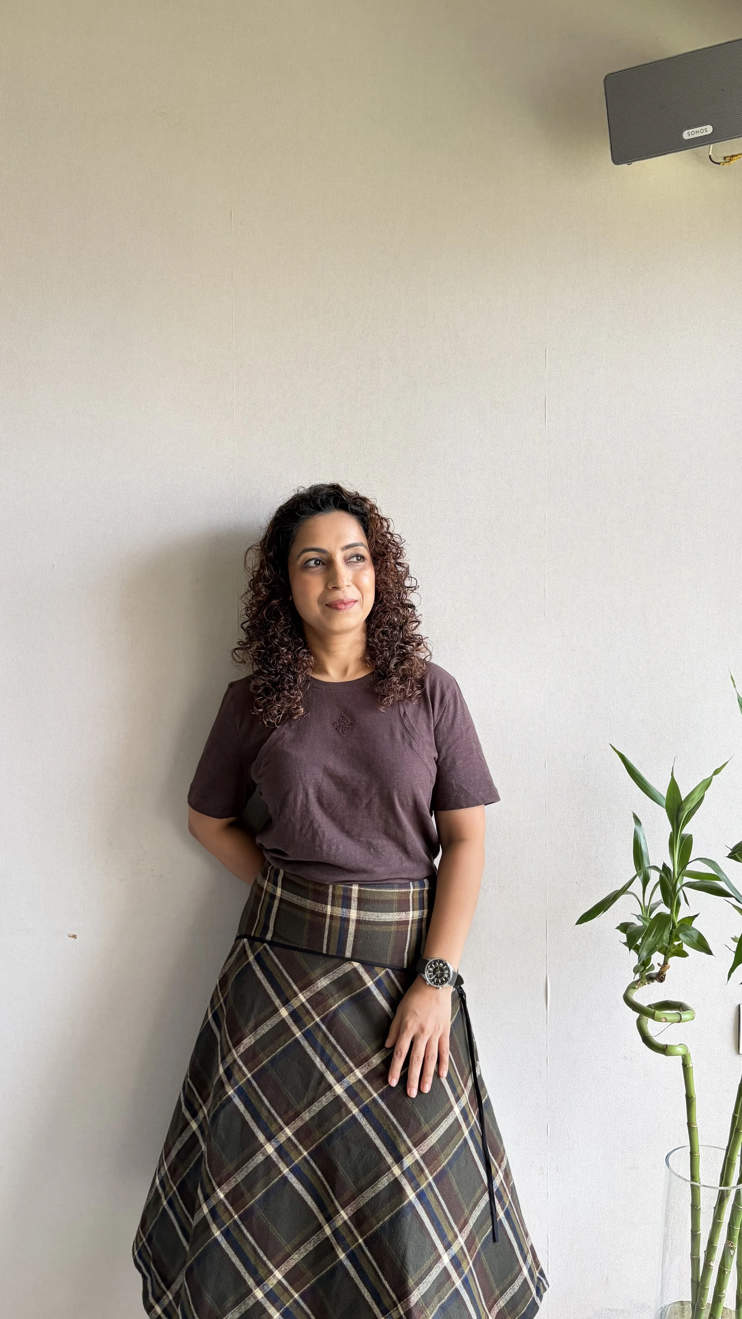 Woman with curly hair wearing a brown t-shirt and plaid skirt stands against a plain wall, next to a tall plant and a wall-mounted speaker.