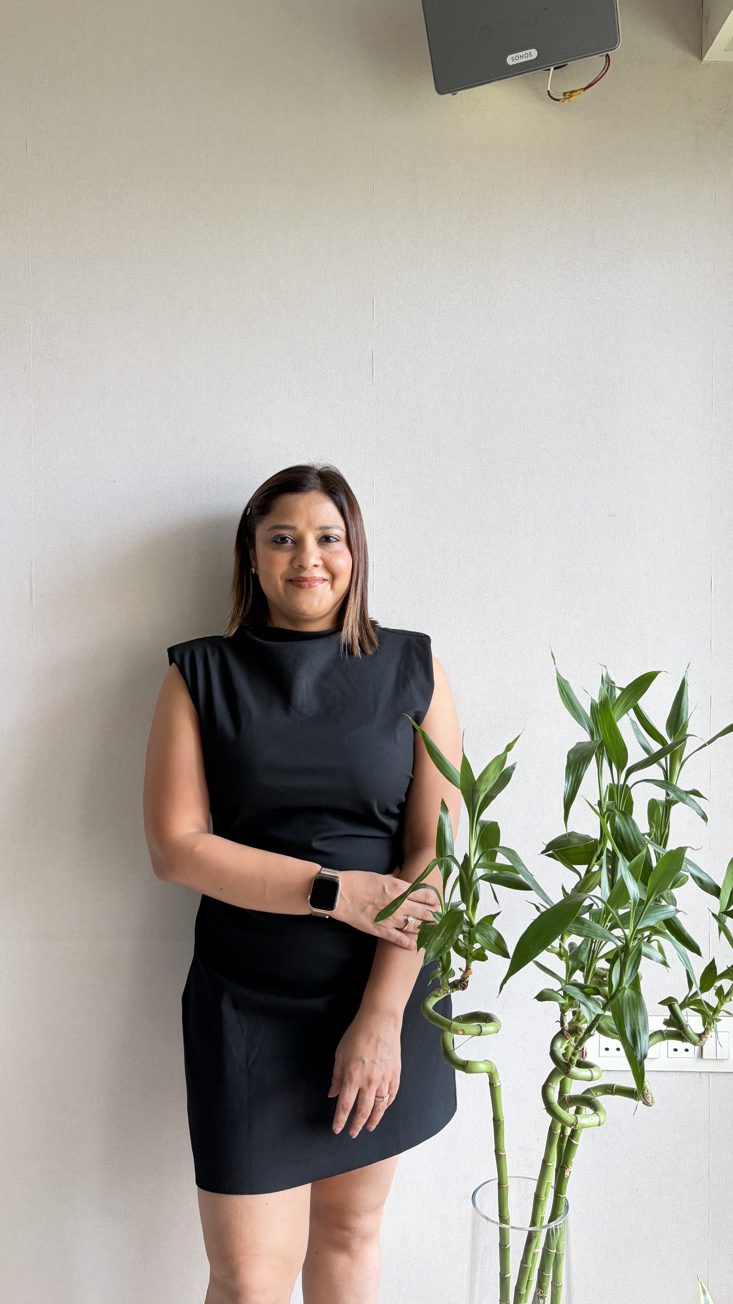 A woman in a black dress standing next to a tall green plant against a plain light-colored wall.