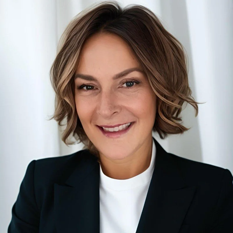 A woman with short, wavy brown hair, wearing a black blazer and a white top, smiling in front of a light-colored background.