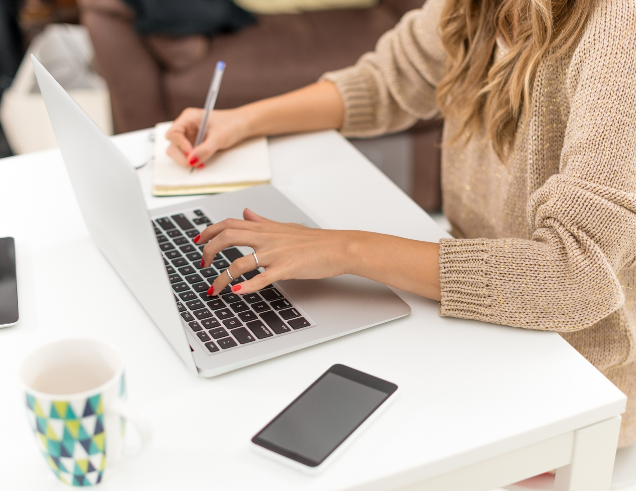 Woman working on a silver laptop at a white desk, taking notes in a small notebook with a pen, with a smartphone and a colorful mug nearby.